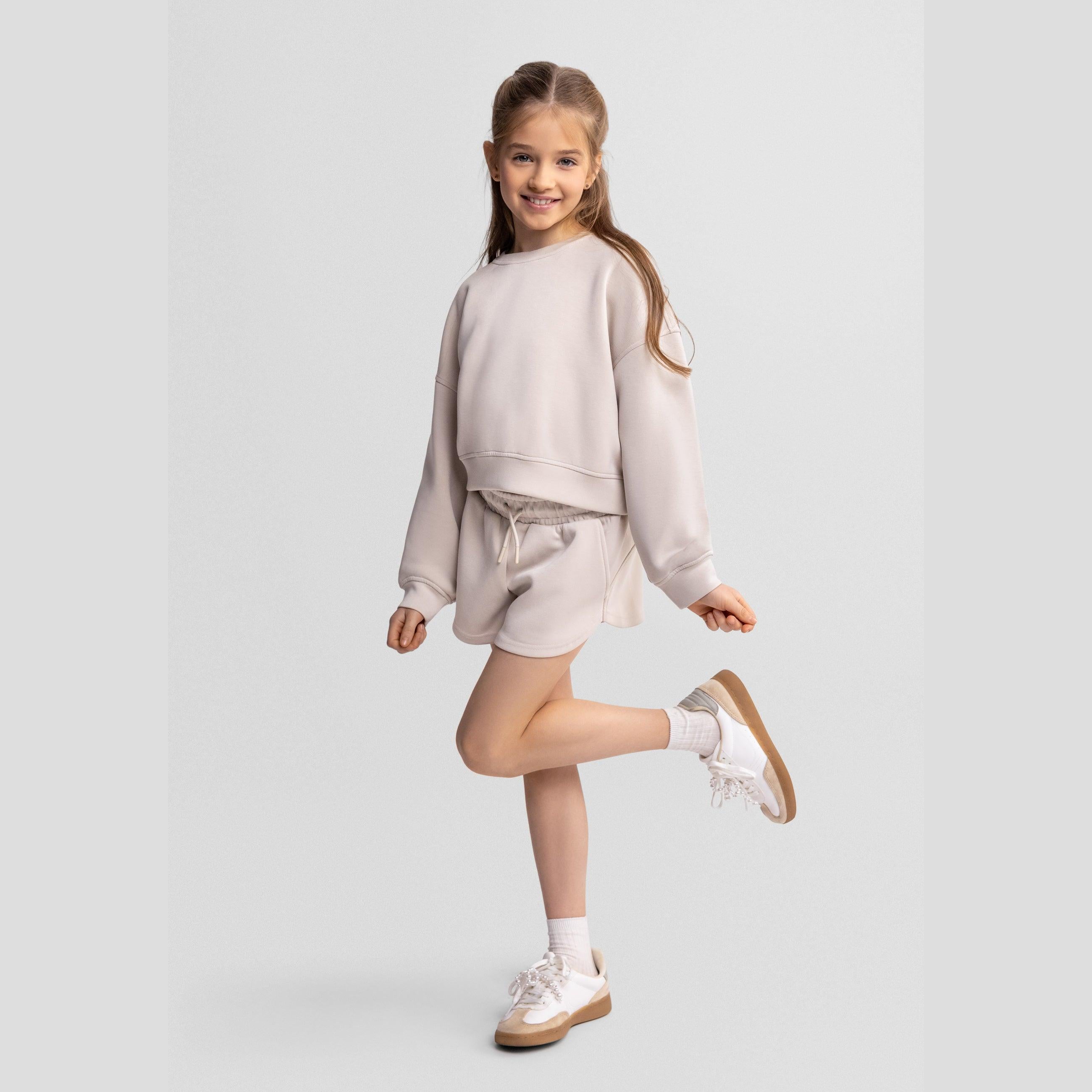 Young girl wearing beige sweatshirt and shorts set, posing playfully on one leg against light grey background