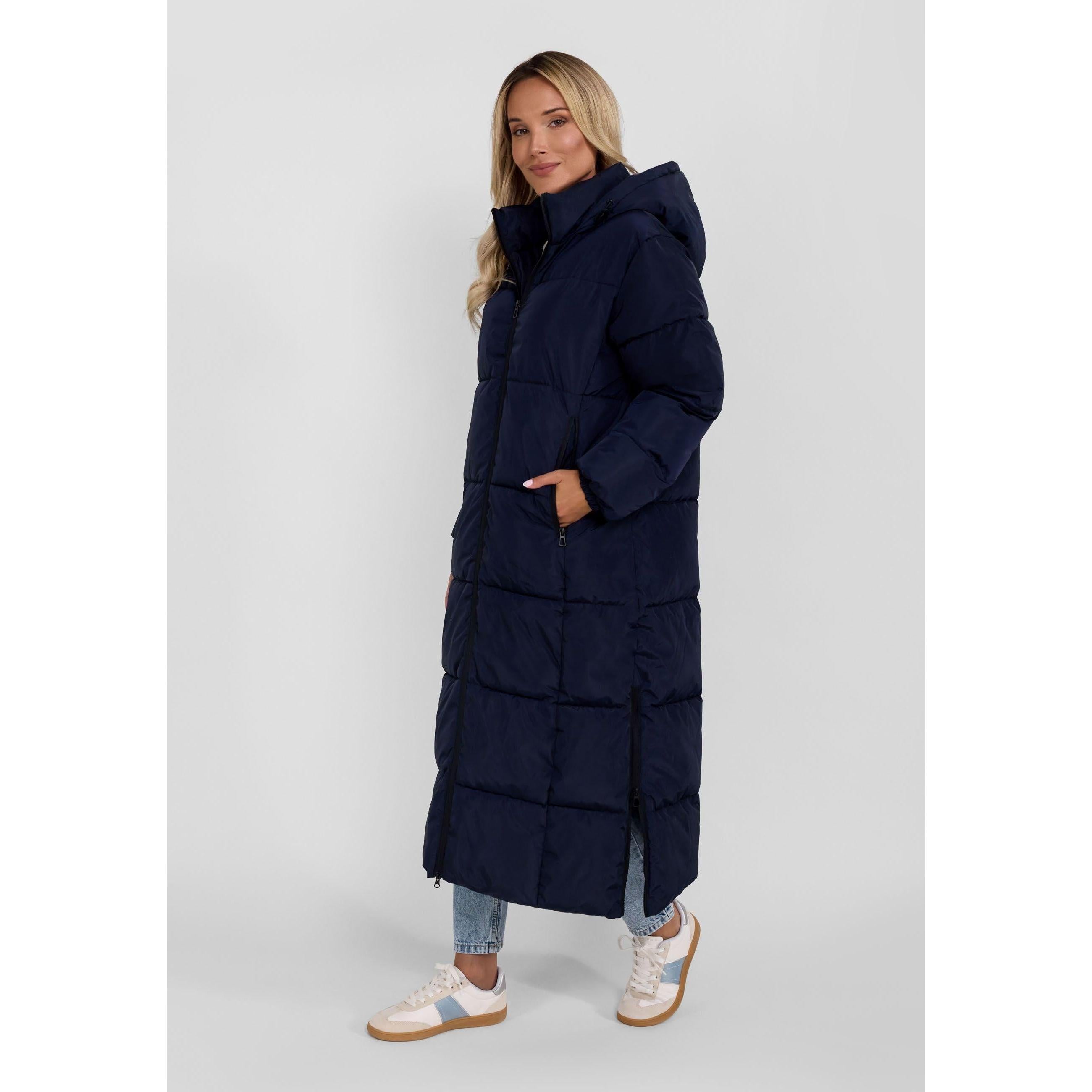 Woman wearing navy long quilted jacket with drawstring hood, hands in pockets, styled with jeans and white sneakers against light background