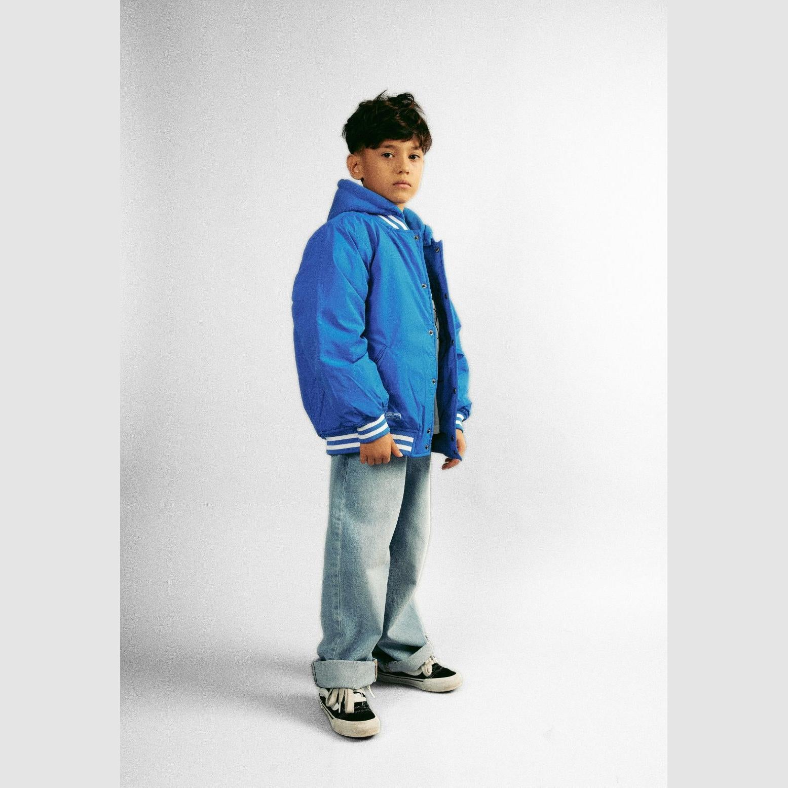 Young boy wearing cobalt blue baseball jacket with detachable hood and light wash jeans, standing against white background