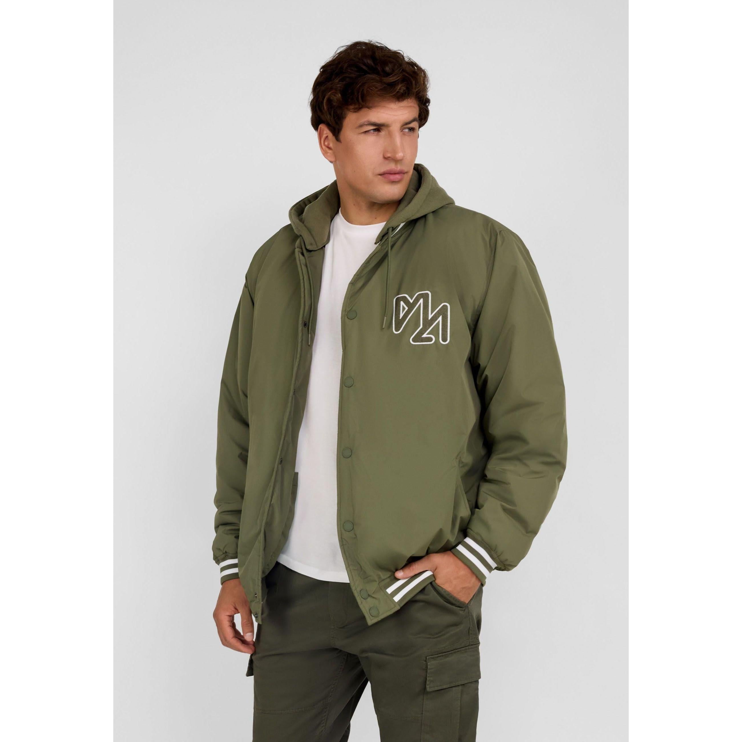 Male model wearing khaki bomber jacket with detachable hood over white t-shirt, standing against grey background
