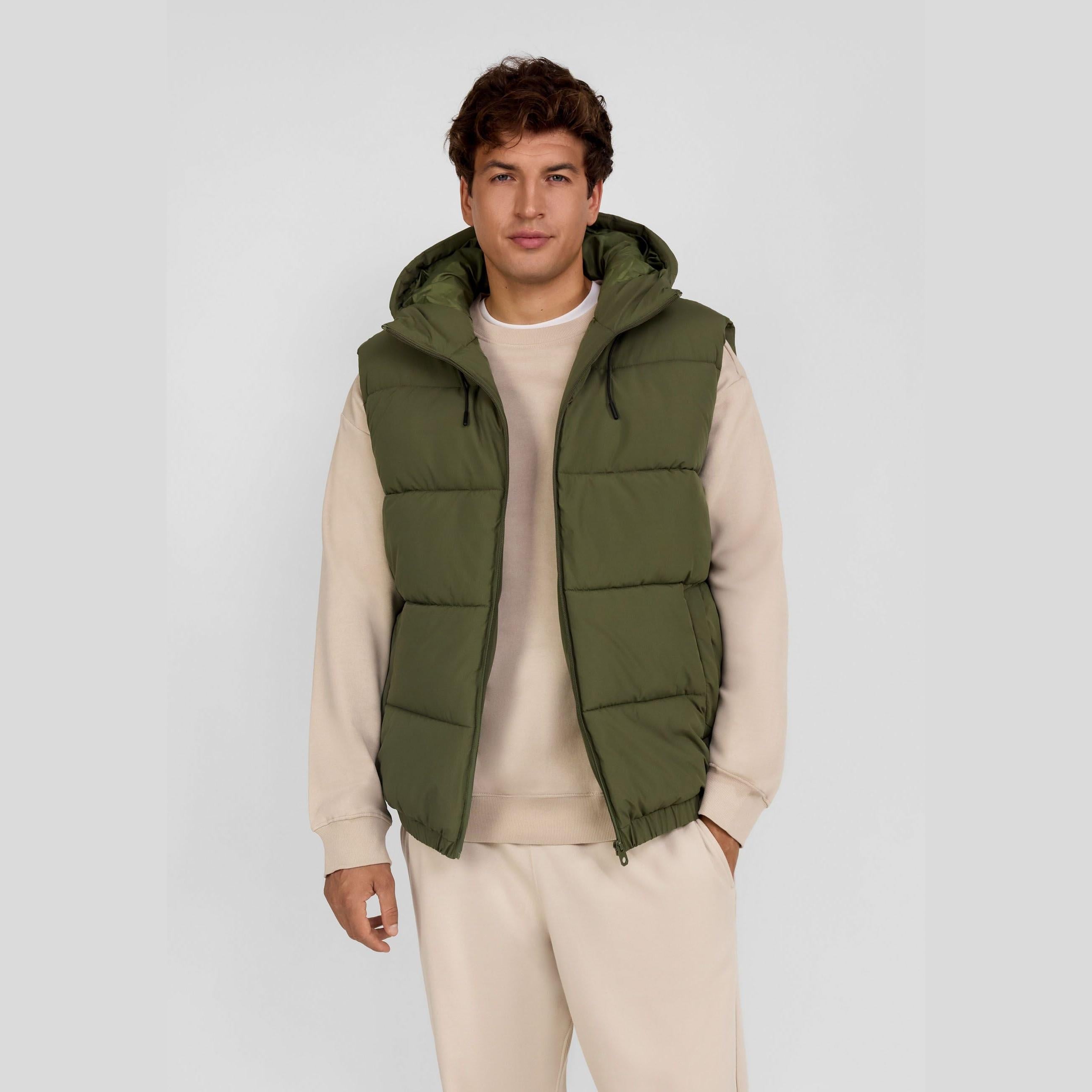 Young male model wearing khaki padded gilet over cream hoodie and joggers, standing against white background, front view showing zip fastening and hood