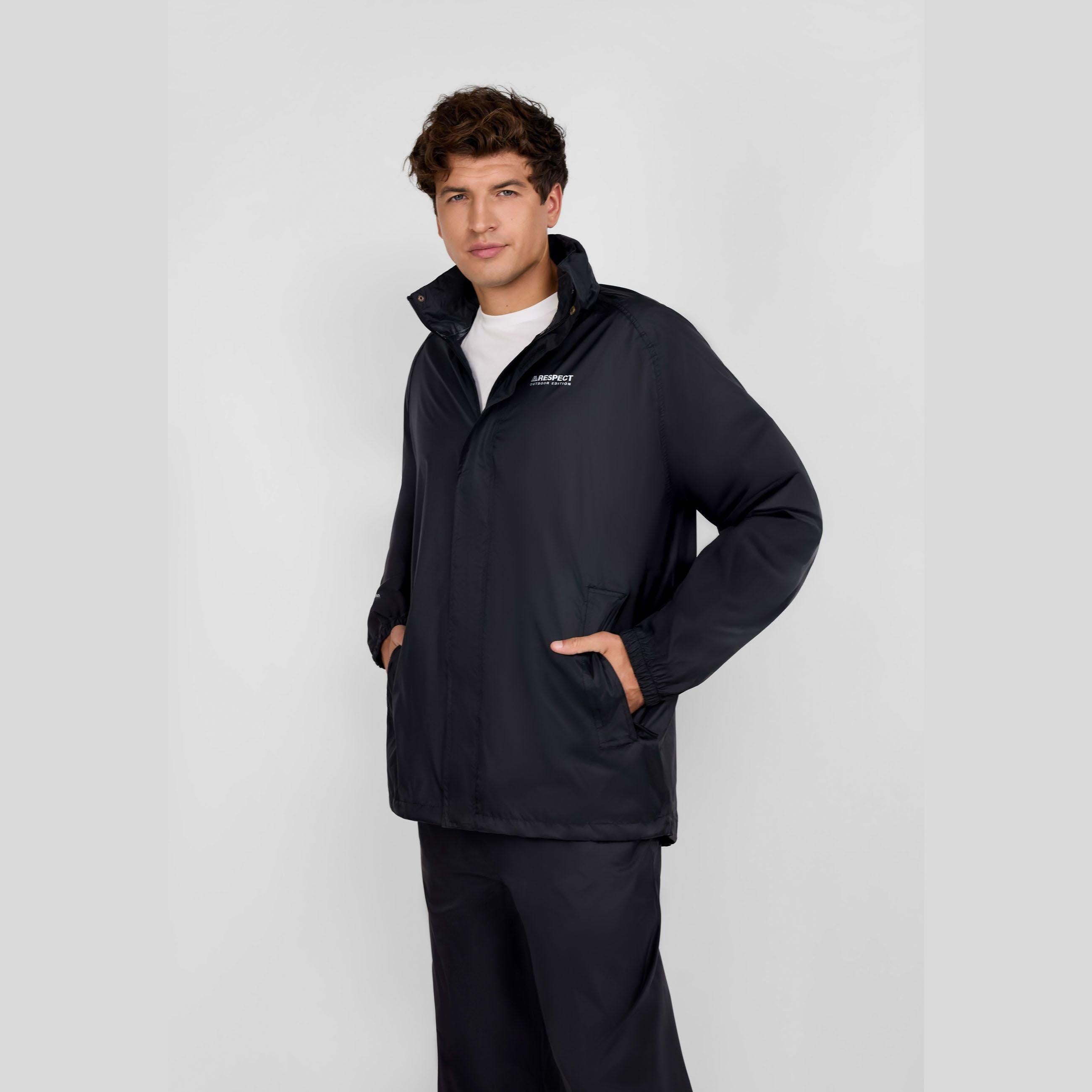 Young man wearing black packaway waterproof jacket with hood, hands in pockets, paired with dark trousers on grey background