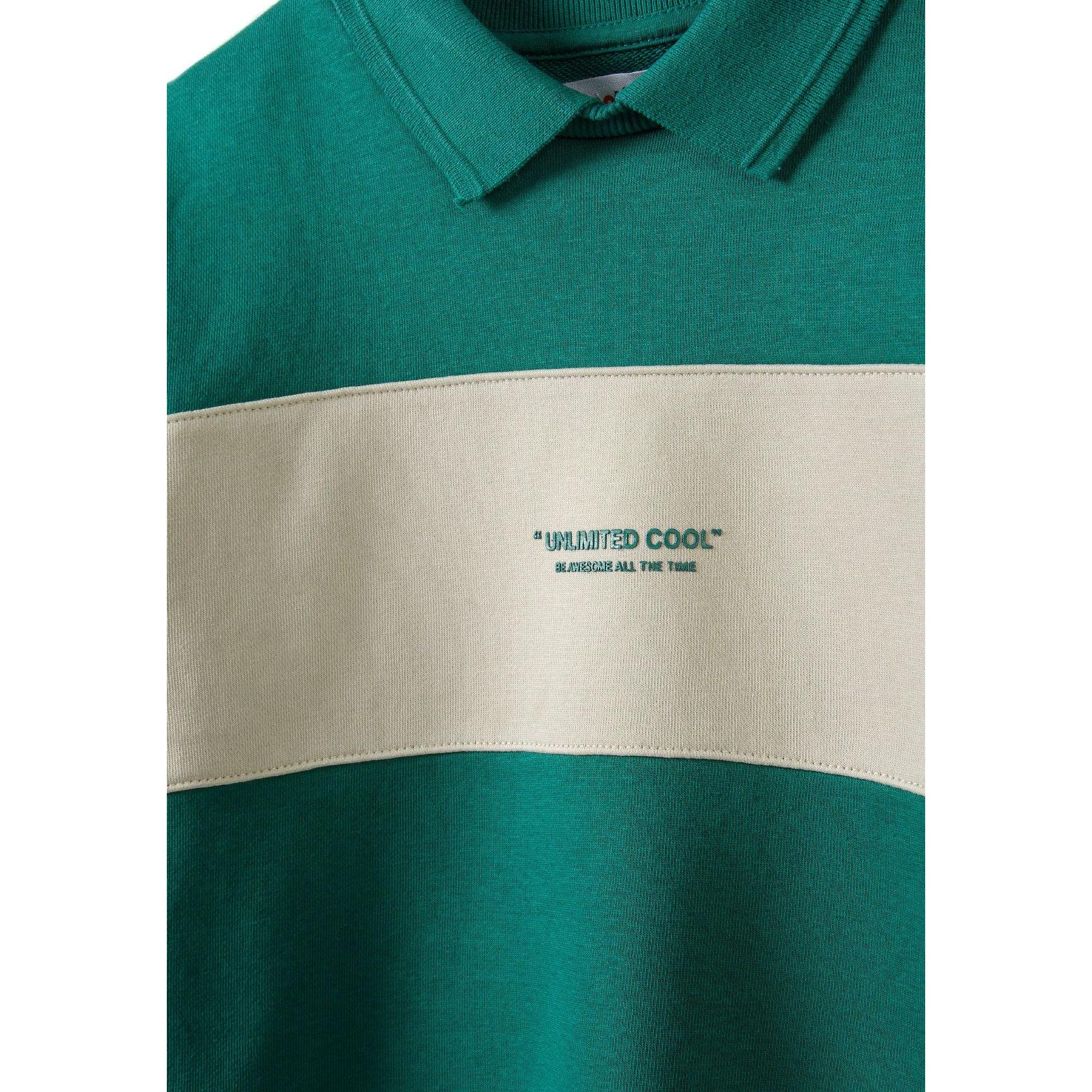 Close-up detail of green fleece polo jumper showing polo collar, cream contrast panel with 'UNLIMITED COOL' text print, and soft fabric texture