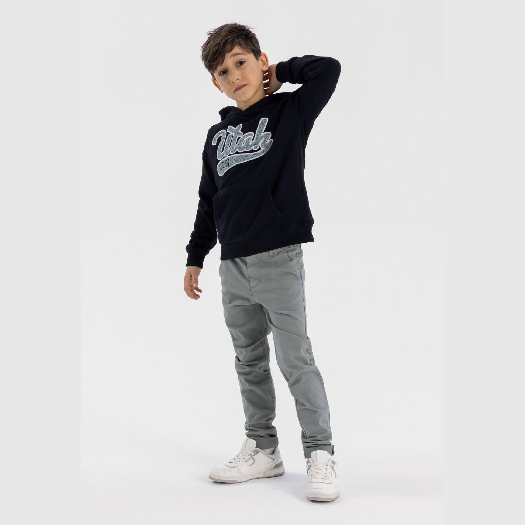 Young boy wearing grey chino trousers with black hoodie and white sneakers, posing against white studio background