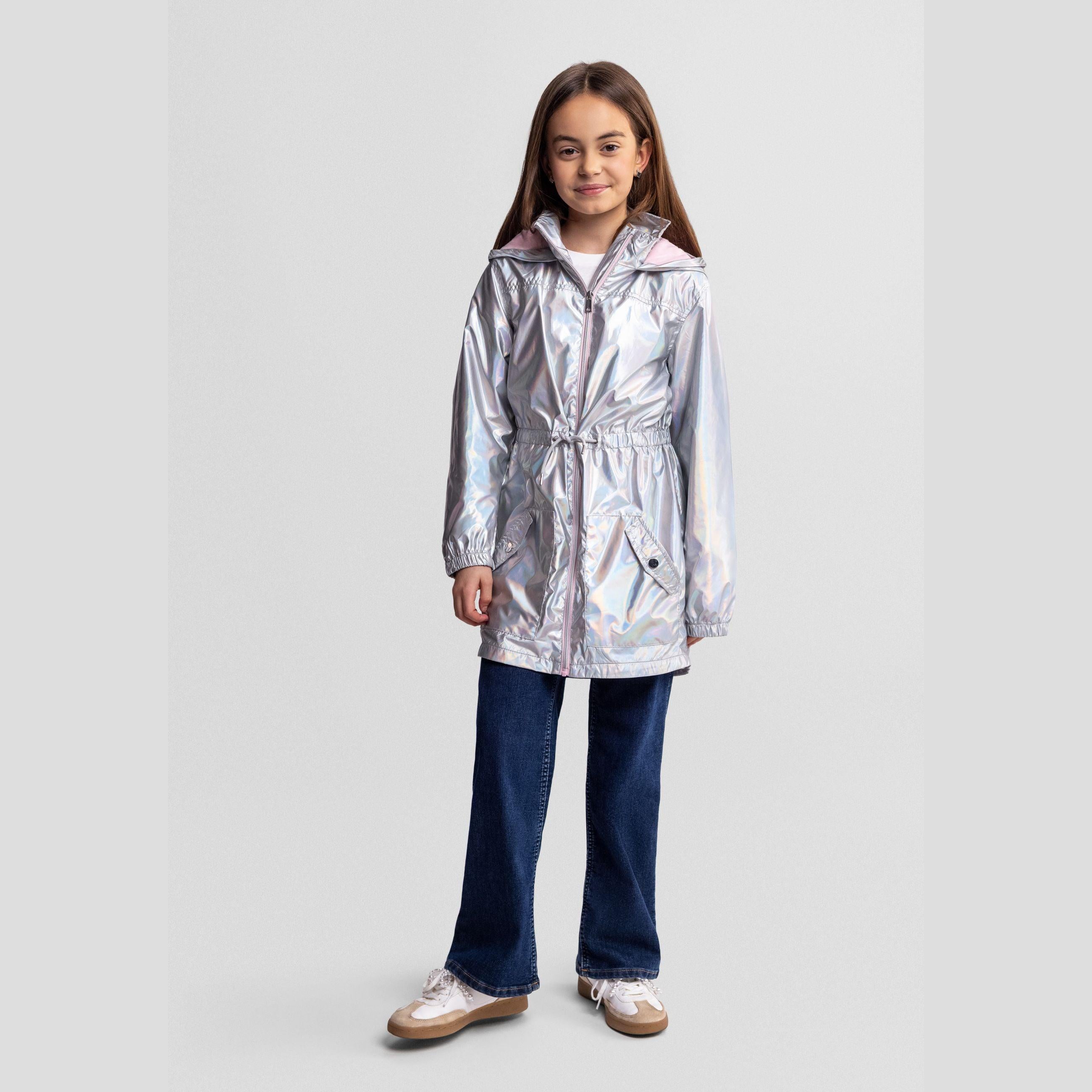 Girl wearing silver iridescent hooded rain jacket with drawstring waist, paired with dark blue jeans and white sneakers, front view on light background