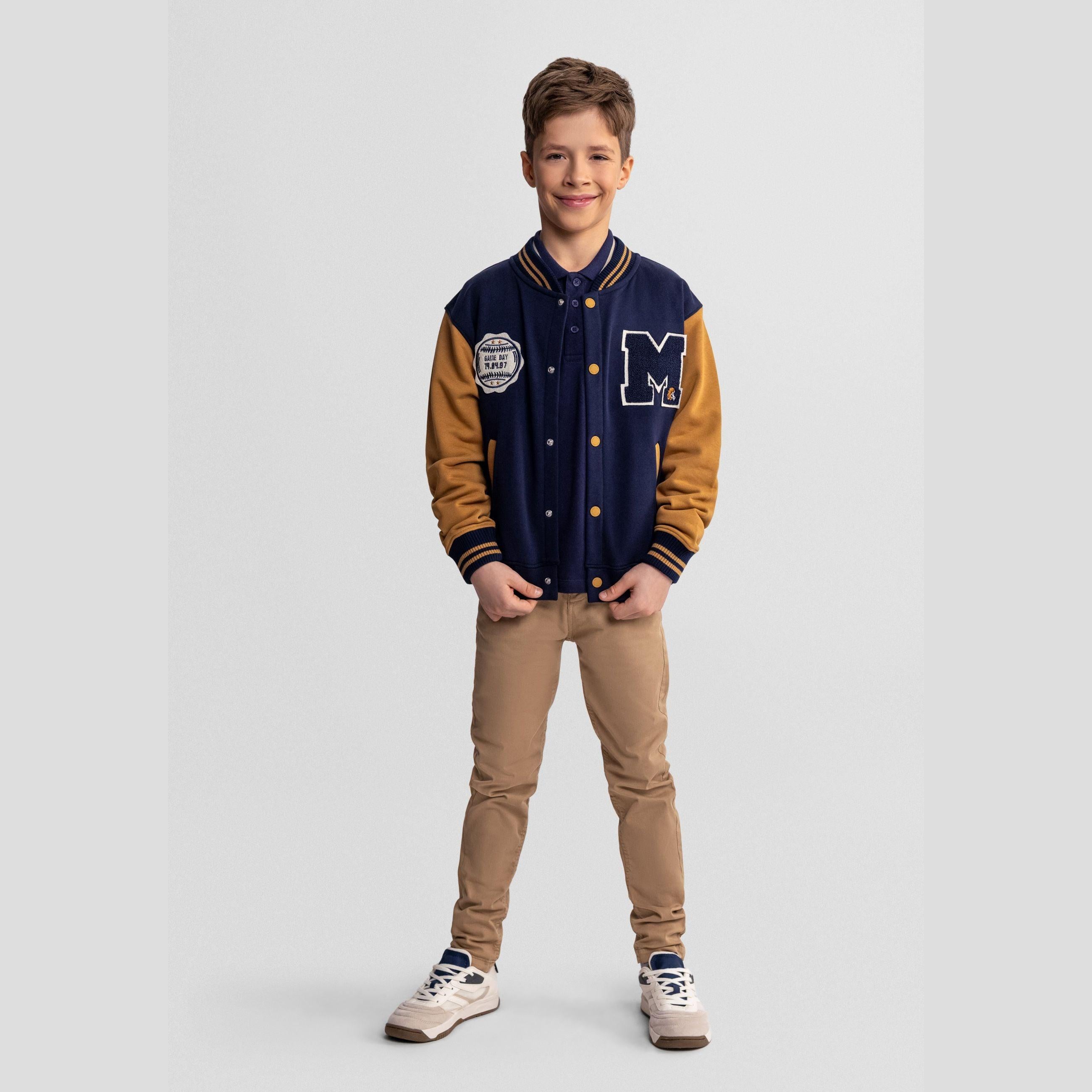 Boy wearing navy baseball jacket with toffee sleeves and beige trousers, standing against white background