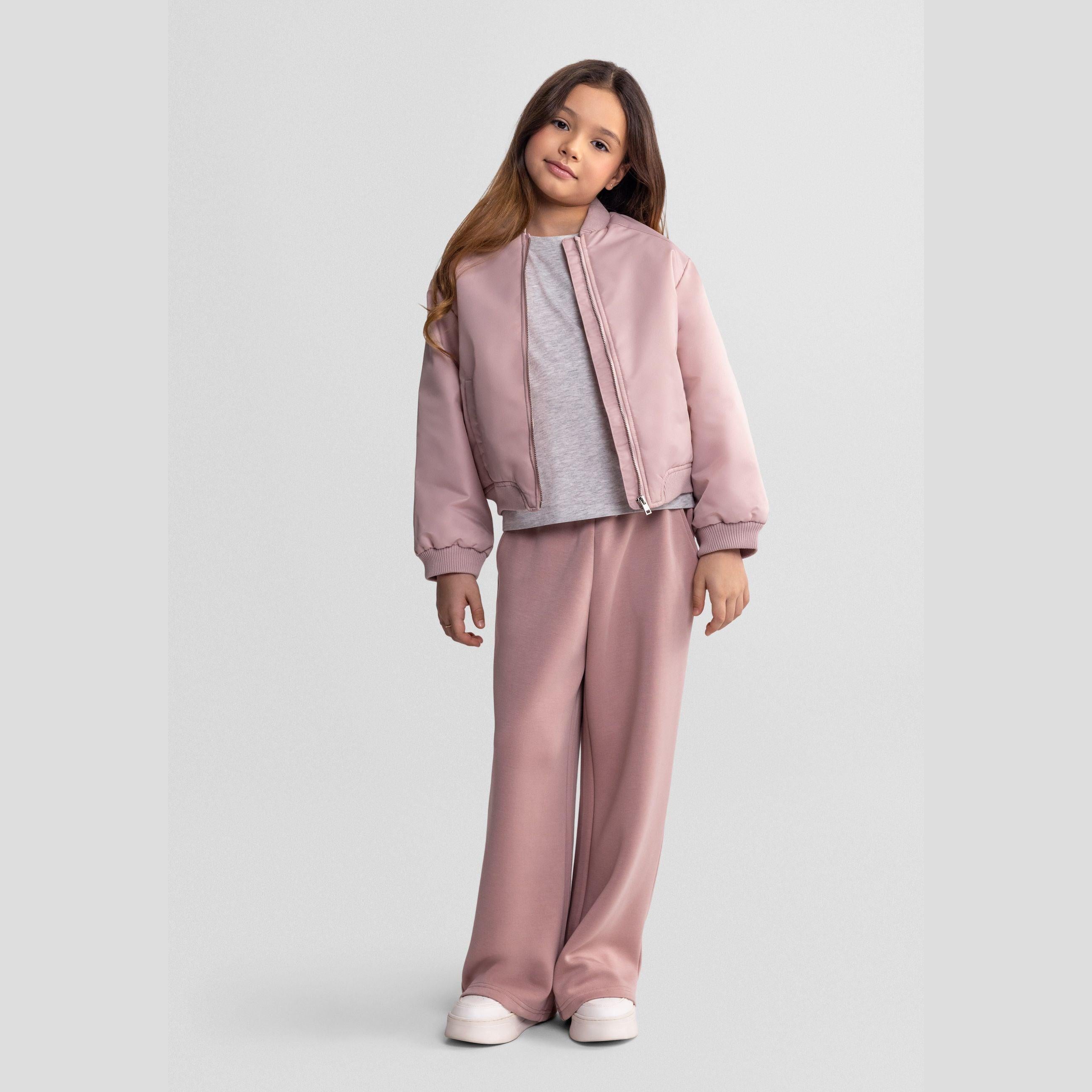 Side view of girl in pink quilted bomber jacket and matching trousers, white sneakers, showing profile against white studio background