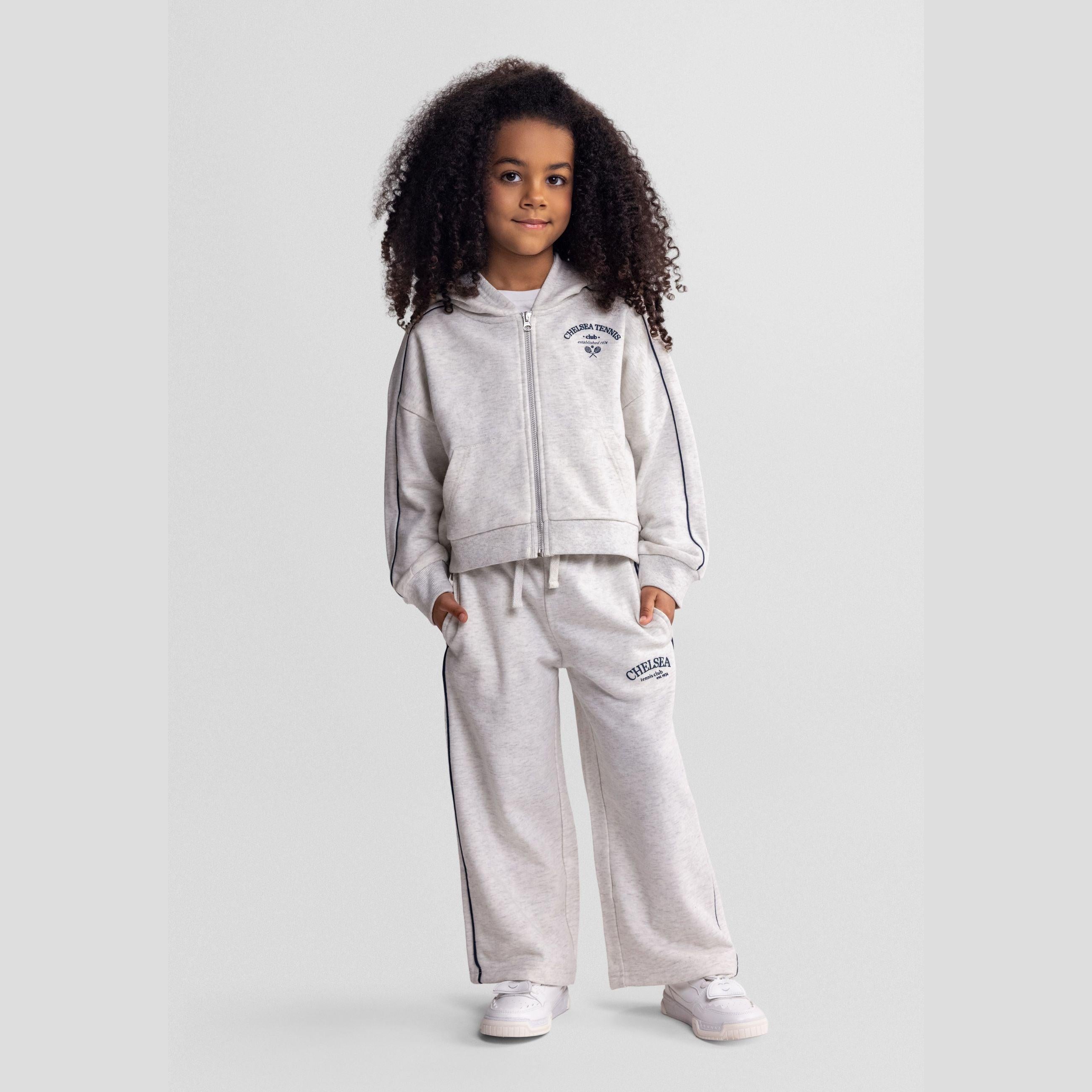 Young girl wearing grey marl hooded tracksuit with Chelsea team club branding, standing against neutral background