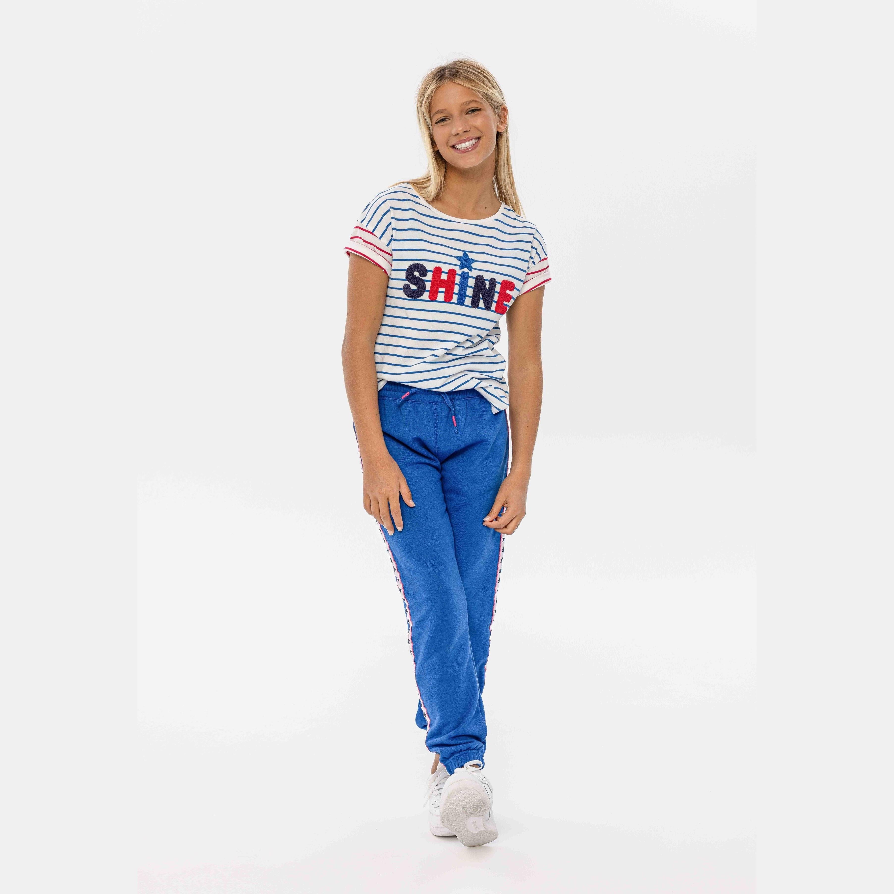 Young girl wearing blue fleece joggers with striped SHINE t-shirt, walking against white background