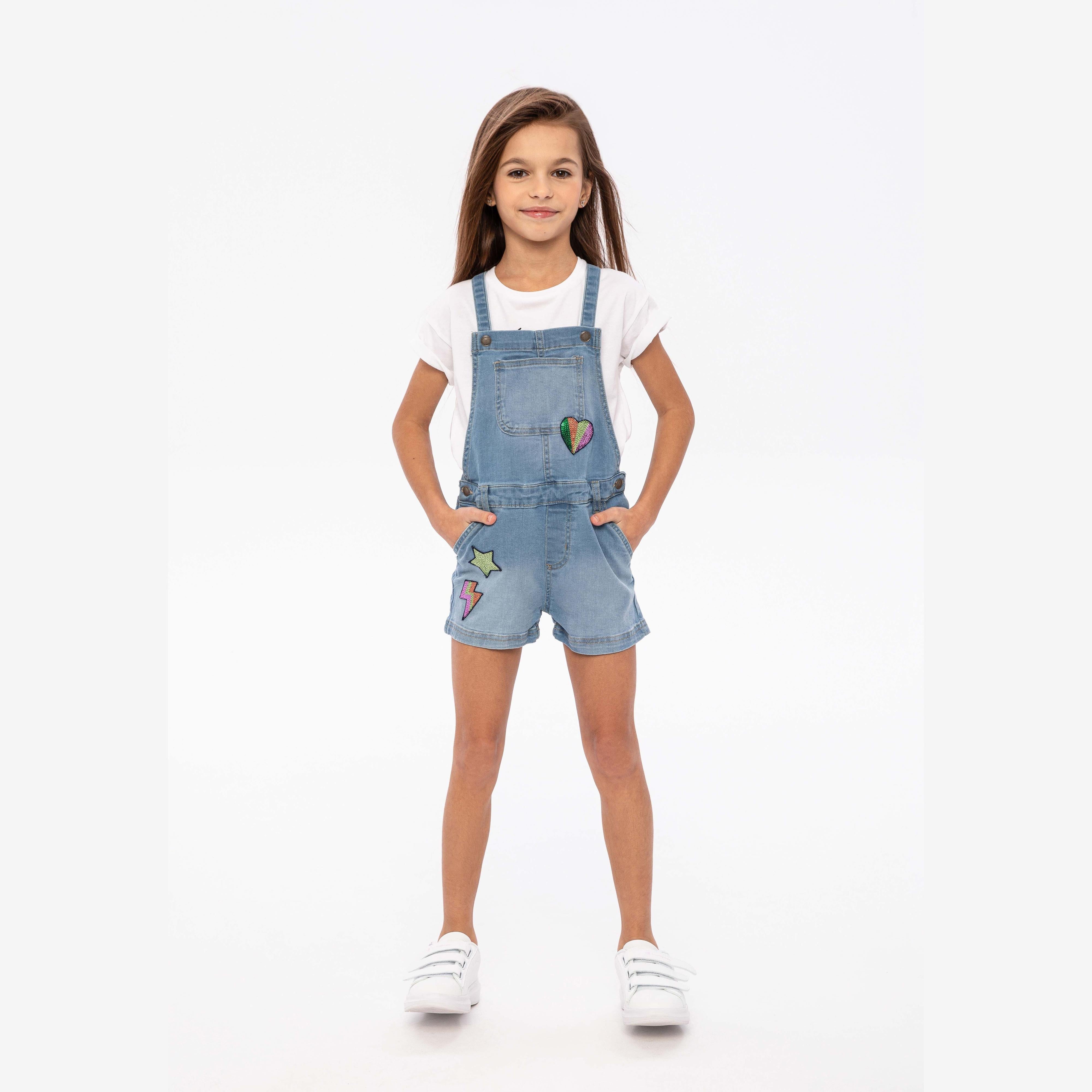 Young girl wearing light blue denim short dungarees with white t-shirt and white sneakers, standing against white studio background
