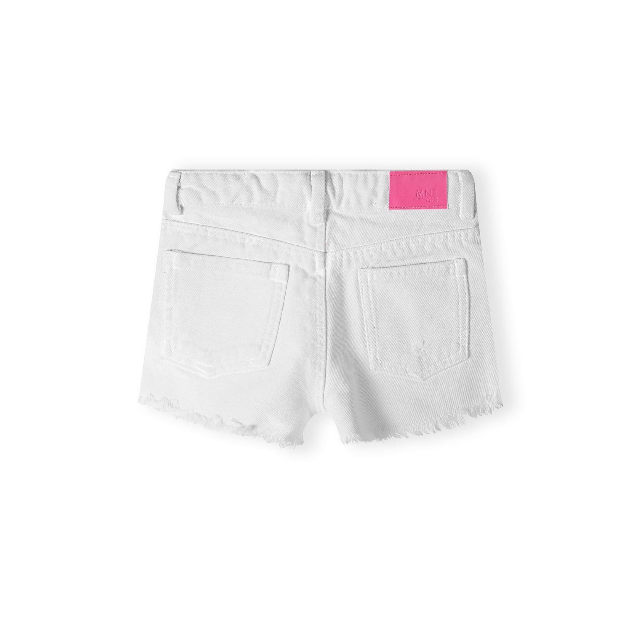 White denim shorts laid flat showing back view with pink Minoti label patch and frayed hem details