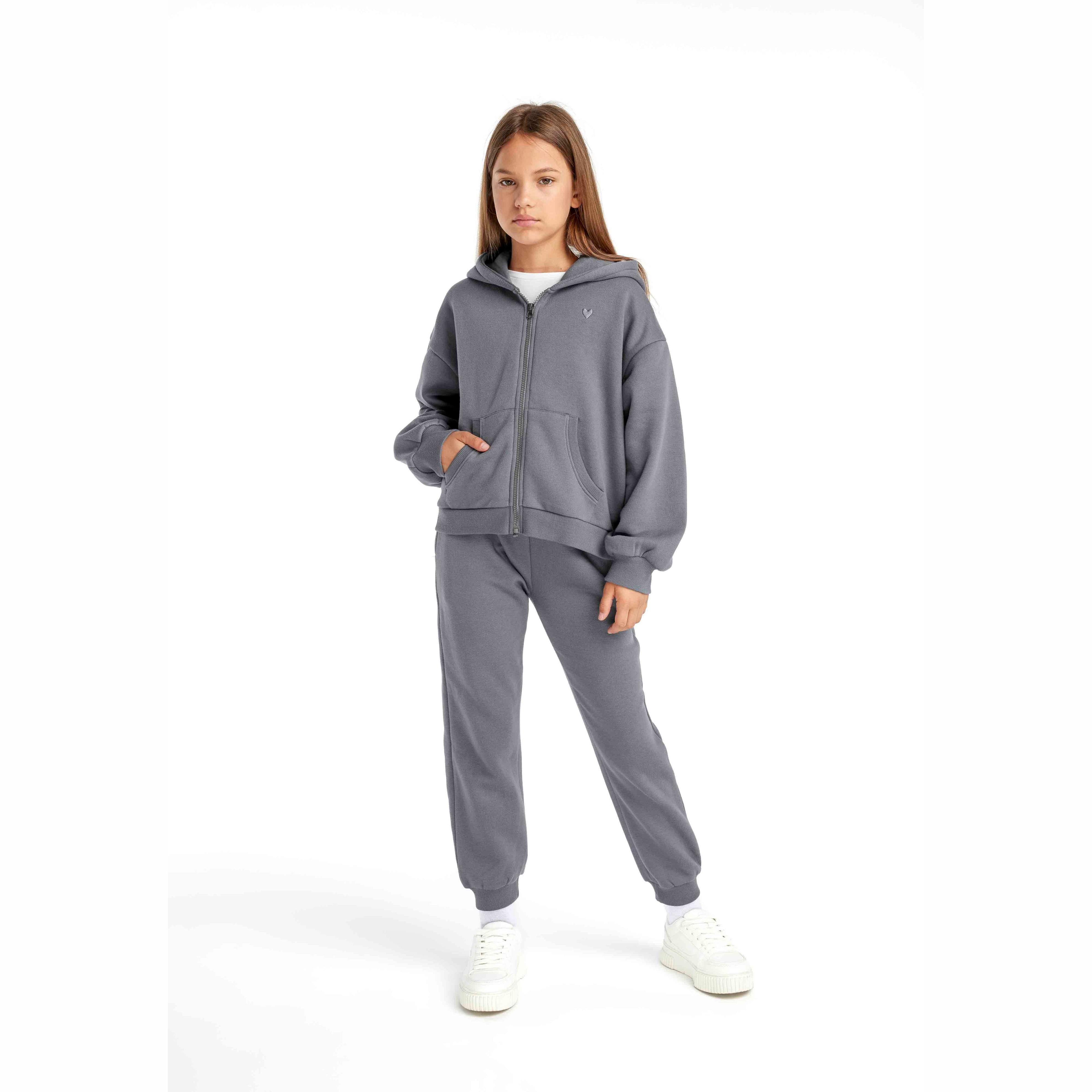 Young girl wearing grey fleece zip hoodie with matching grey joggers and white trainers on white background