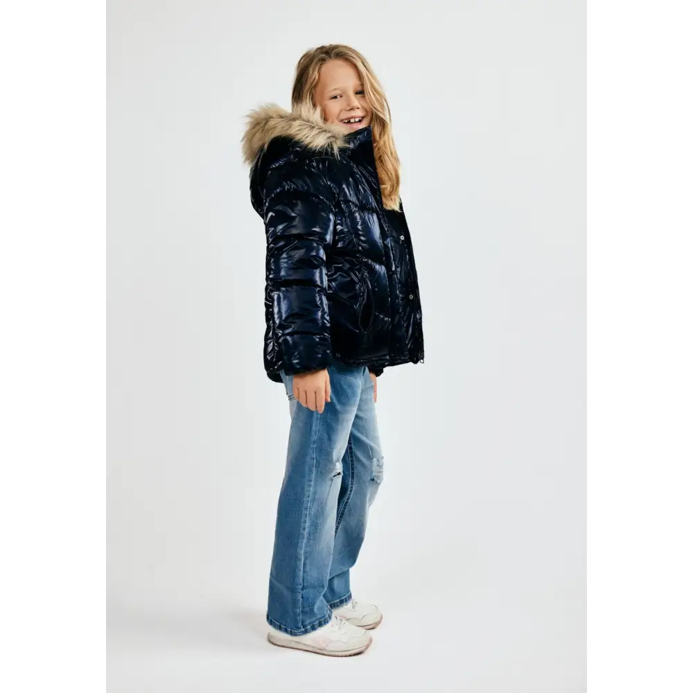 Young girl in navy metallic winter jacket with hood up showing faux fur trim, wearing light blue jeans and white shoes, side angle against white background