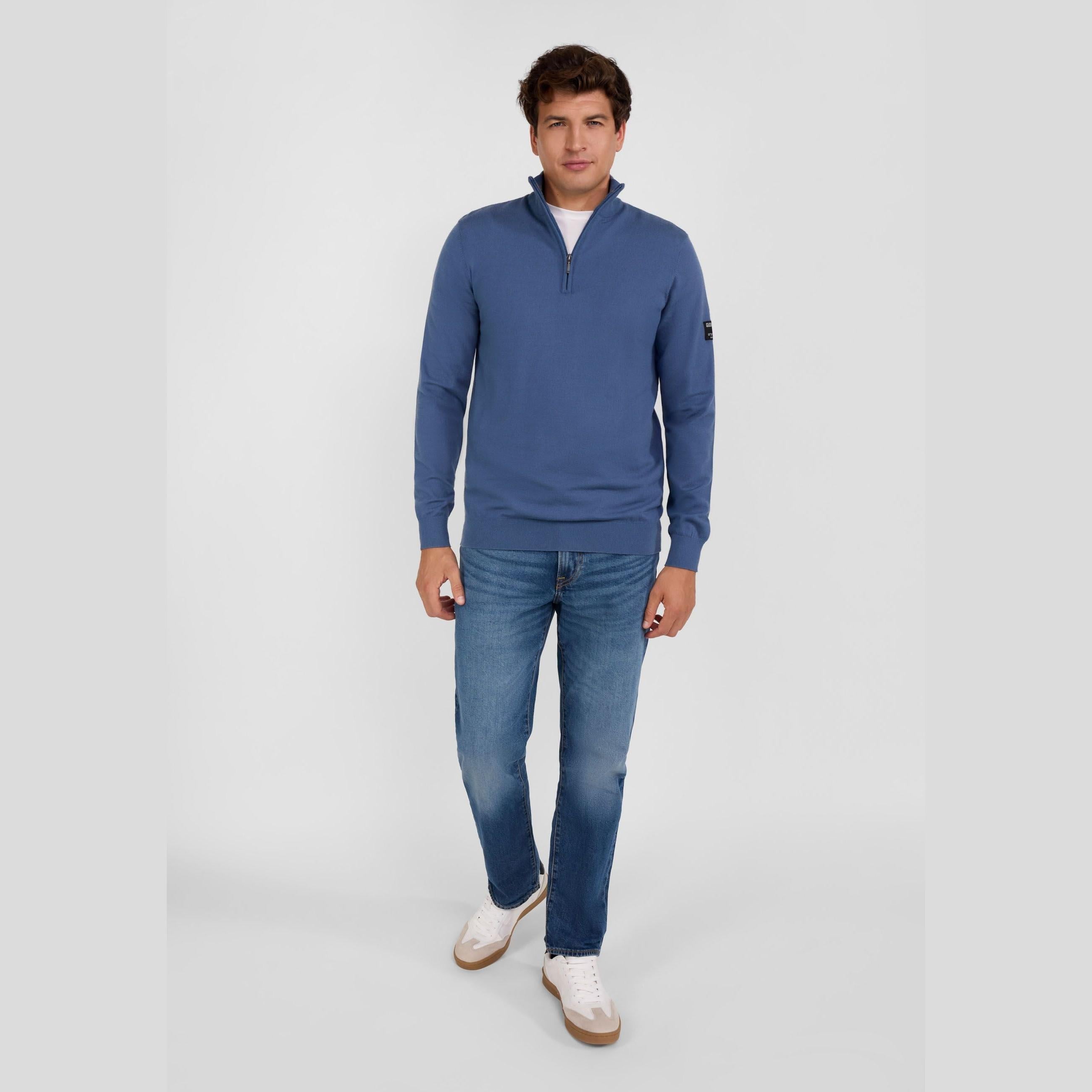 Man in blue half zip jumper shown from side angle, relaxed pose with hands in pockets, white studio background