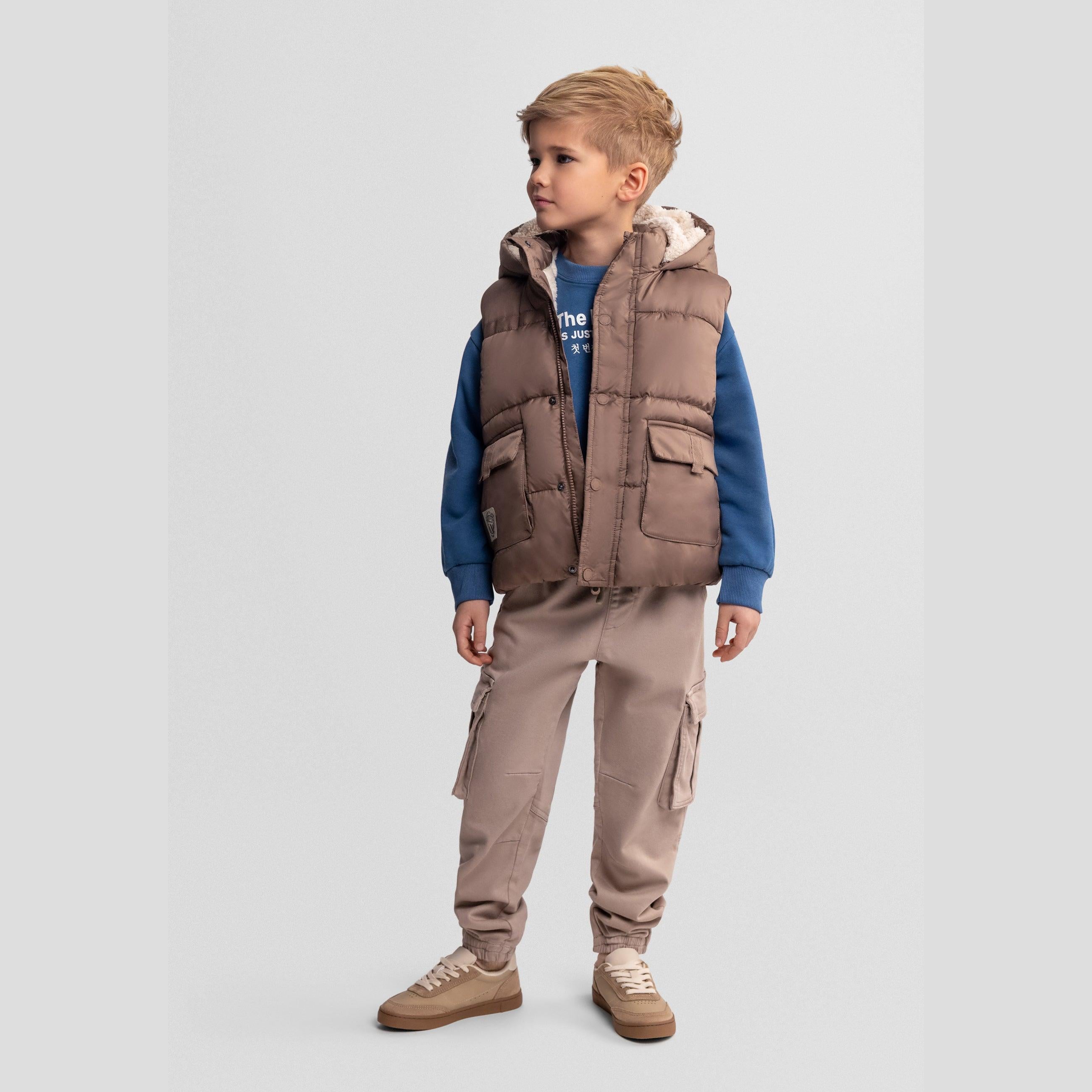 Boy wearing beige quilted hooded gilet over blue sweatshirt with beige cargo trousers and trainers on grey background