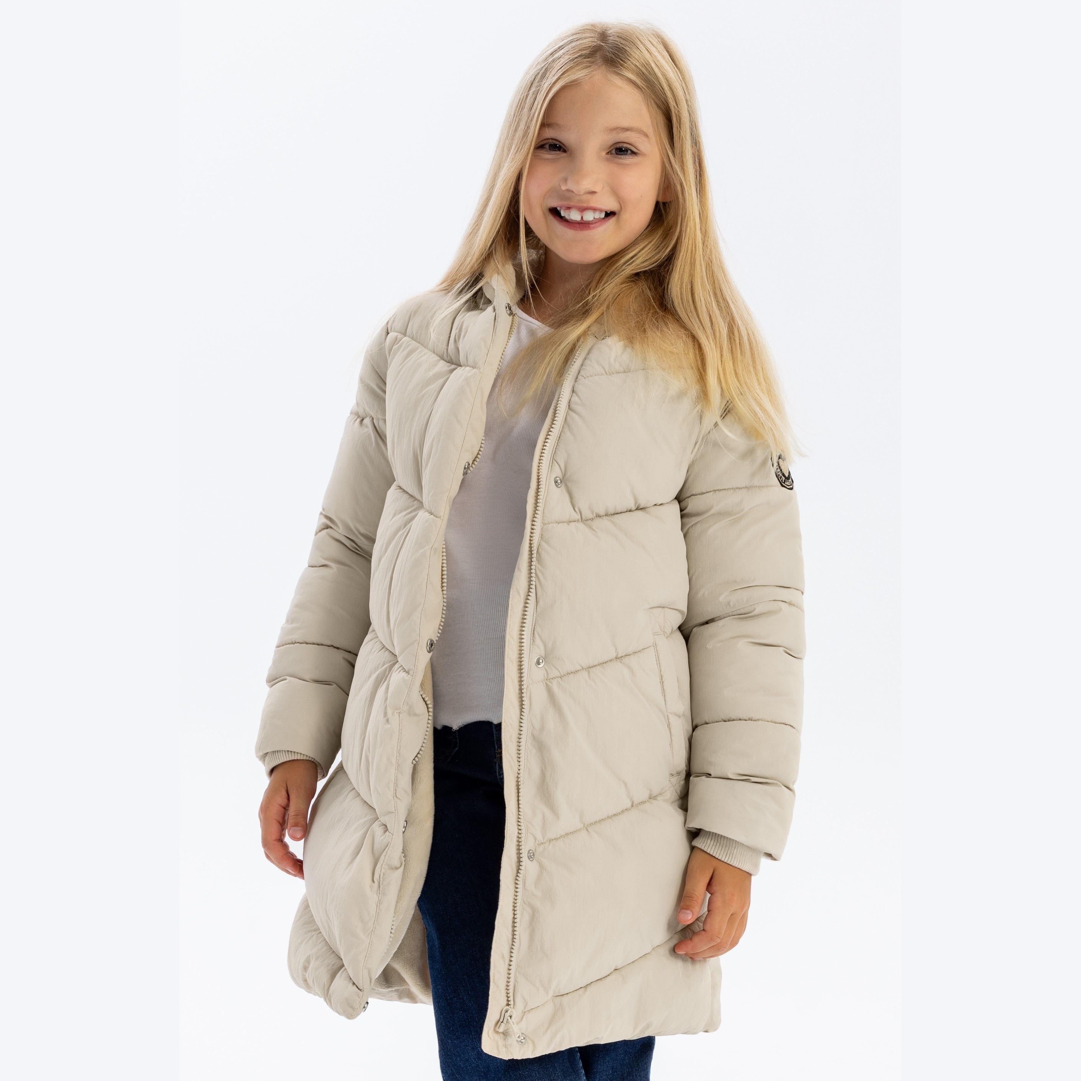 Young blonde girl wearing beige long quilted puffer jacket with hood, smiling at camera against white studio background