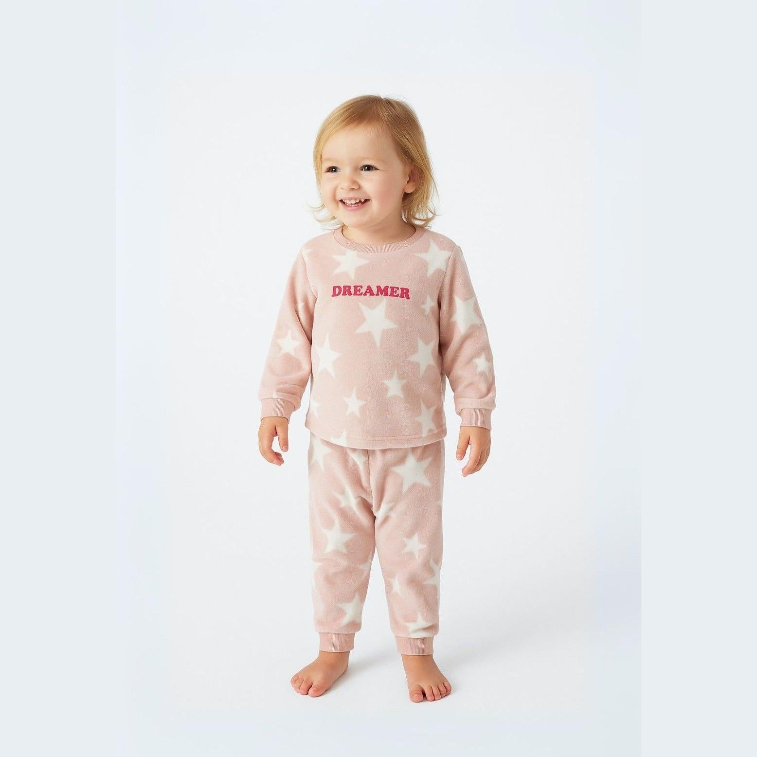 Young girl wearing pink fleece pyjama set with white stars and DREAMER text, smiling against white studio background