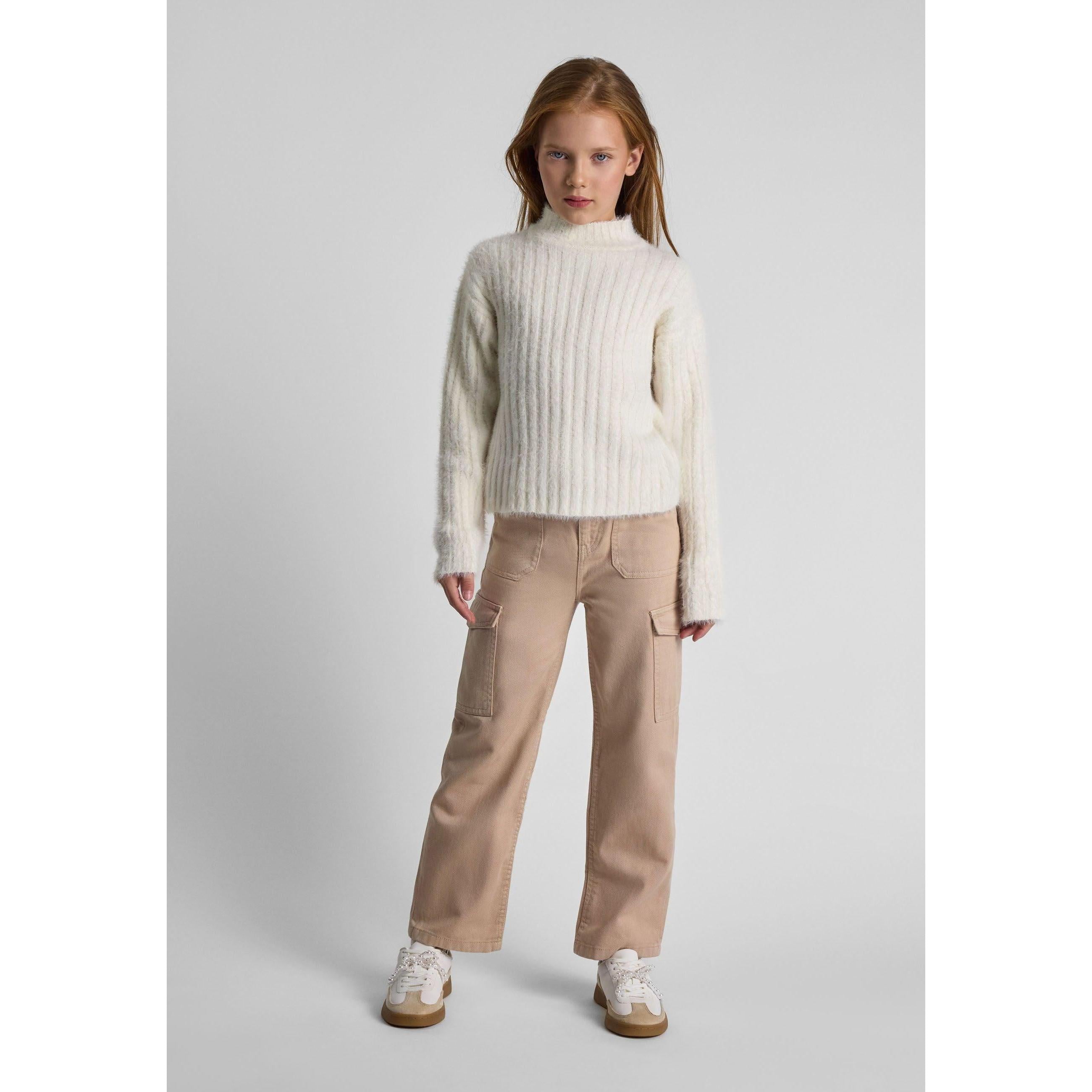 Young girl wearing cream ribbed turtleneck jumper with beige trousers and white trainers, standing against grey background