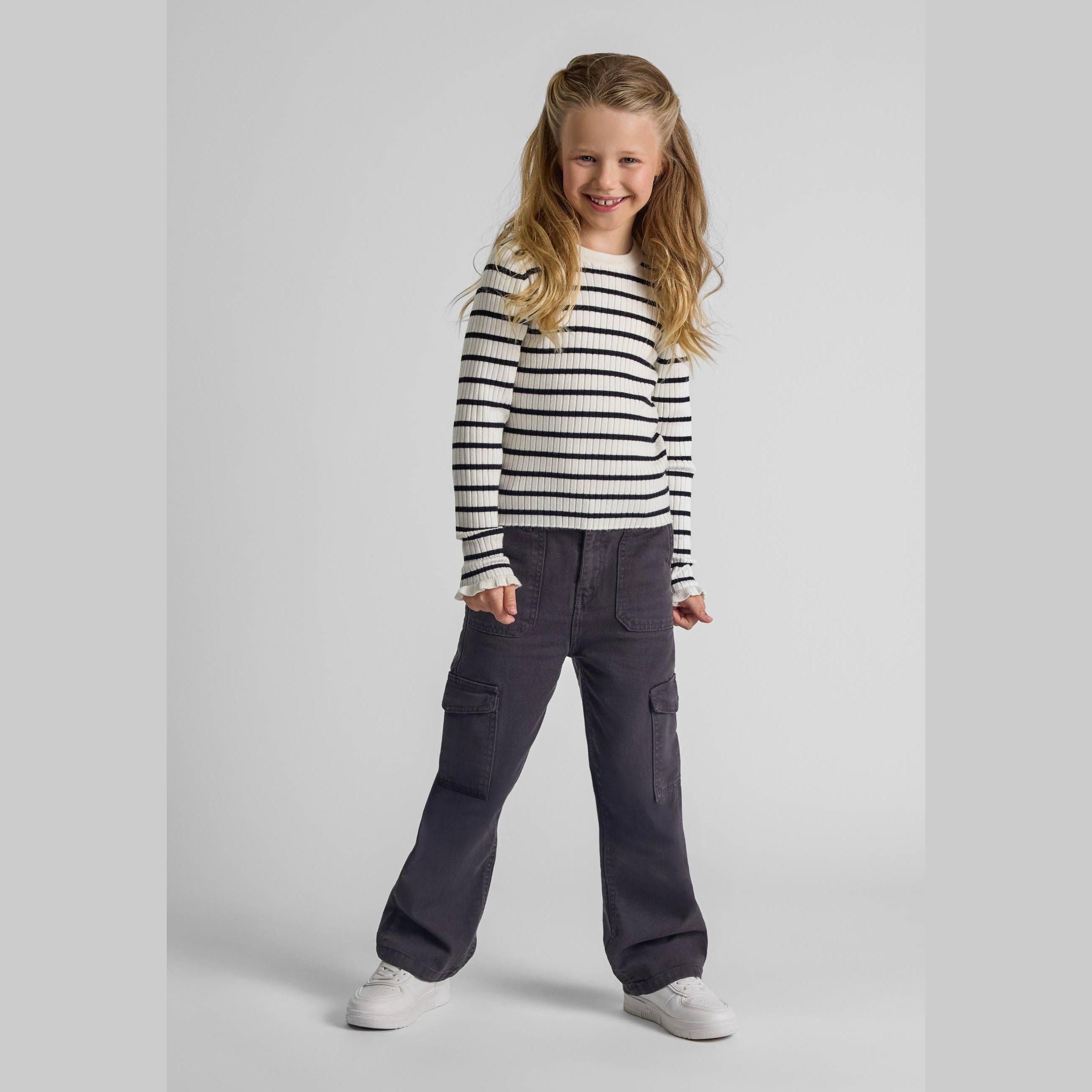 Young blonde girl wearing cream and black striped ribbed jumper with dark cargo trousers, standing against light grey background