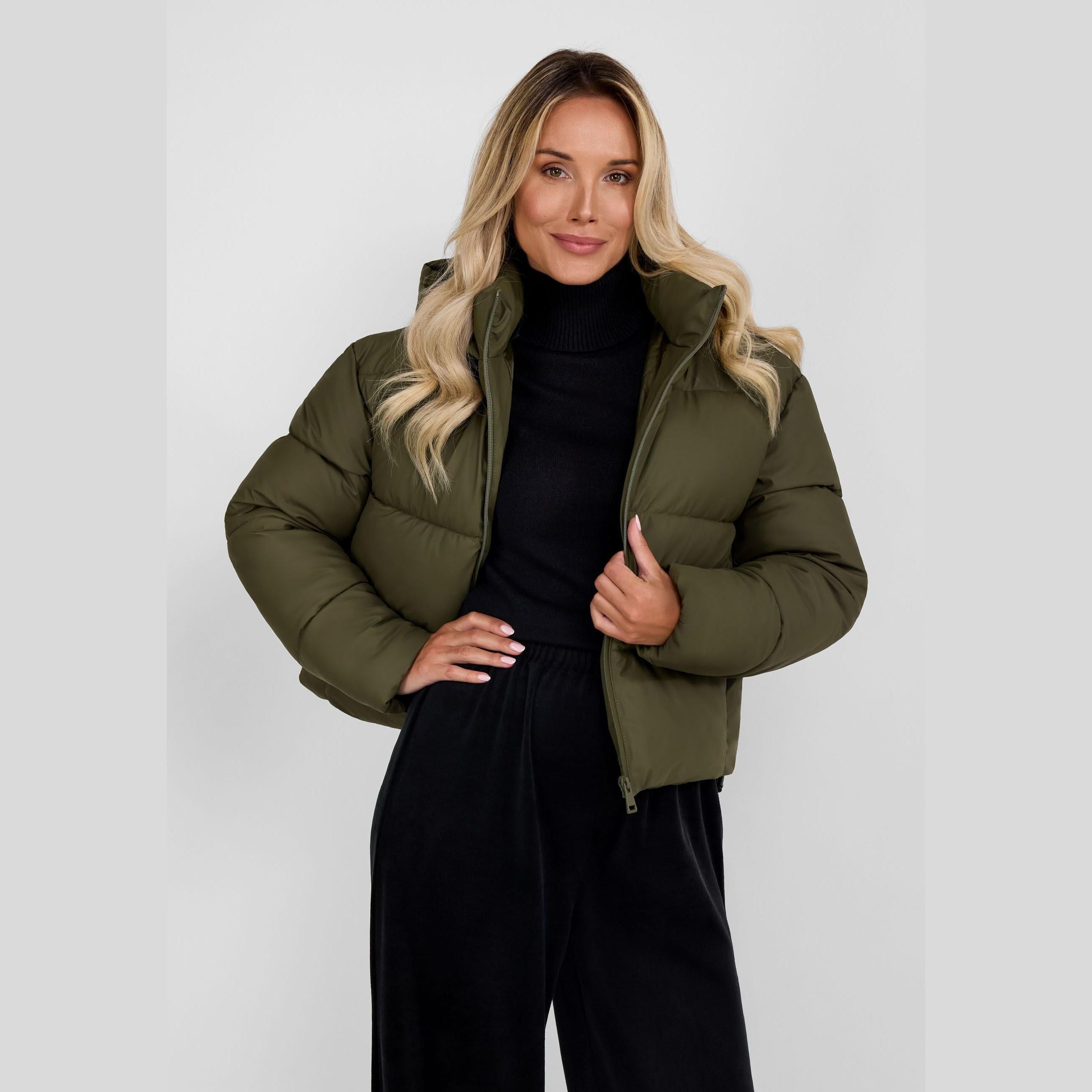 Woman wearing khaki quilted jacket with hood over black turtleneck and trousers, front view on white background