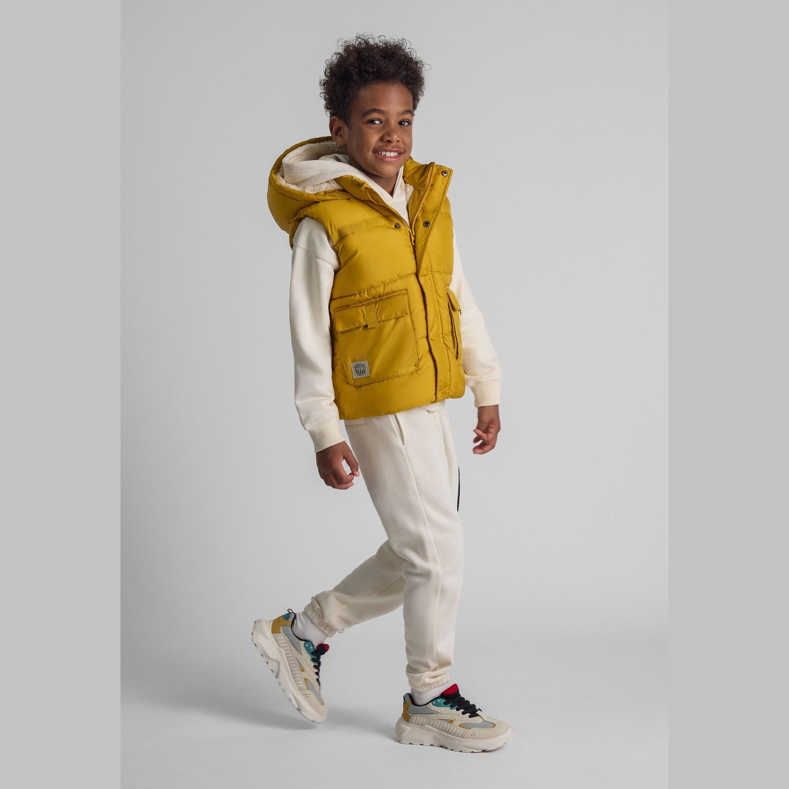 Young child wearing mustard yellow hooded gilet over cream hoodie and joggers, walking pose against grey background