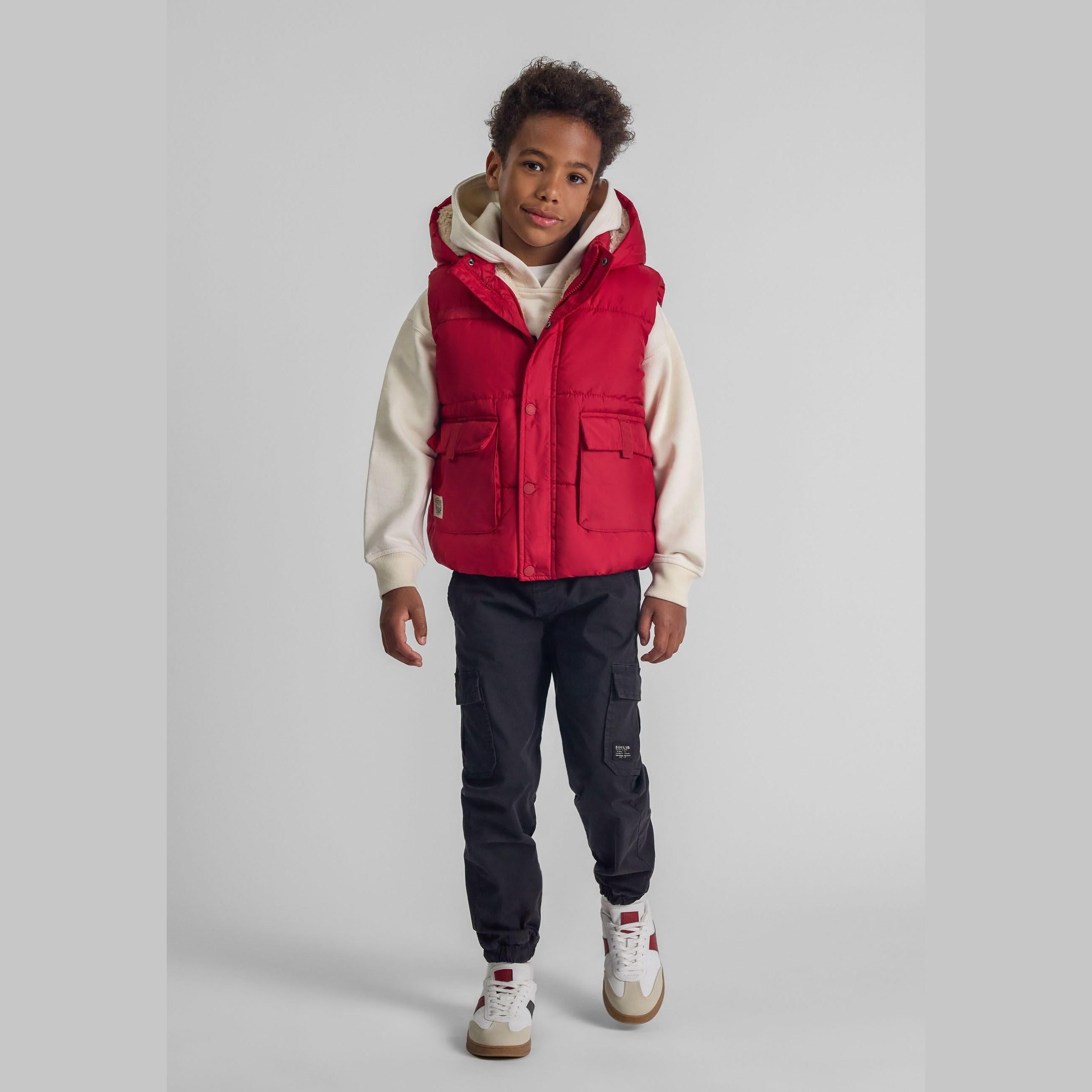 Young boy wearing red quilted gilet over cream hoodie with navy cargo pants against white background