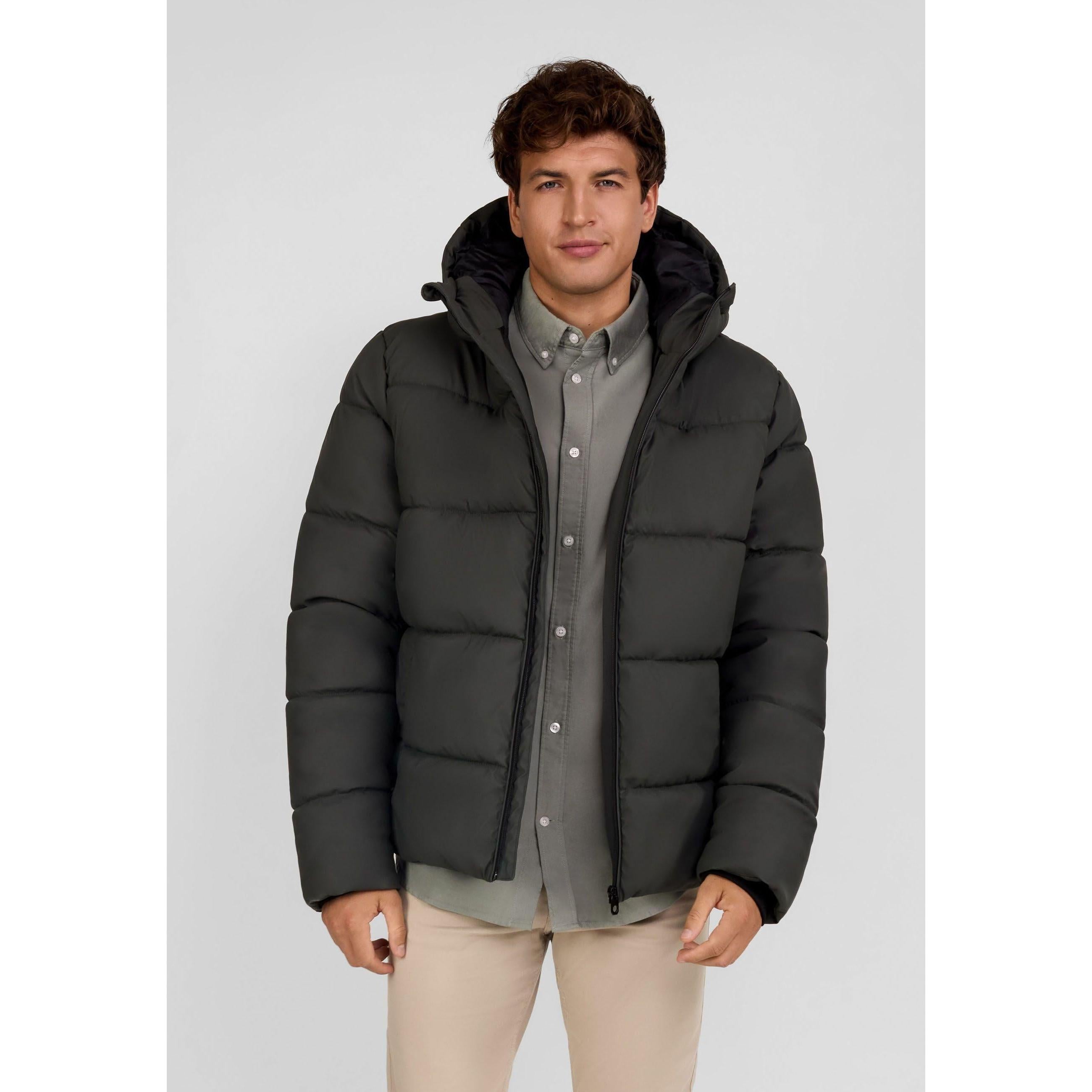 Male model wearing khaki padded jacket with hood over grey shirt and beige trousers against white background