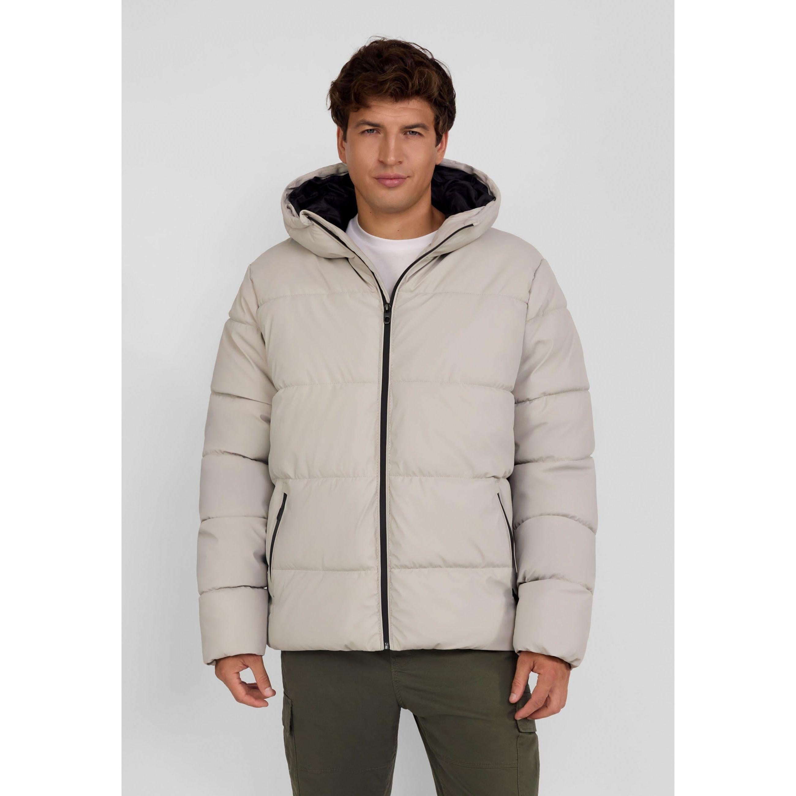 Young male model wearing beige padded jacket with hood up, front zip closed, standing against light grey background