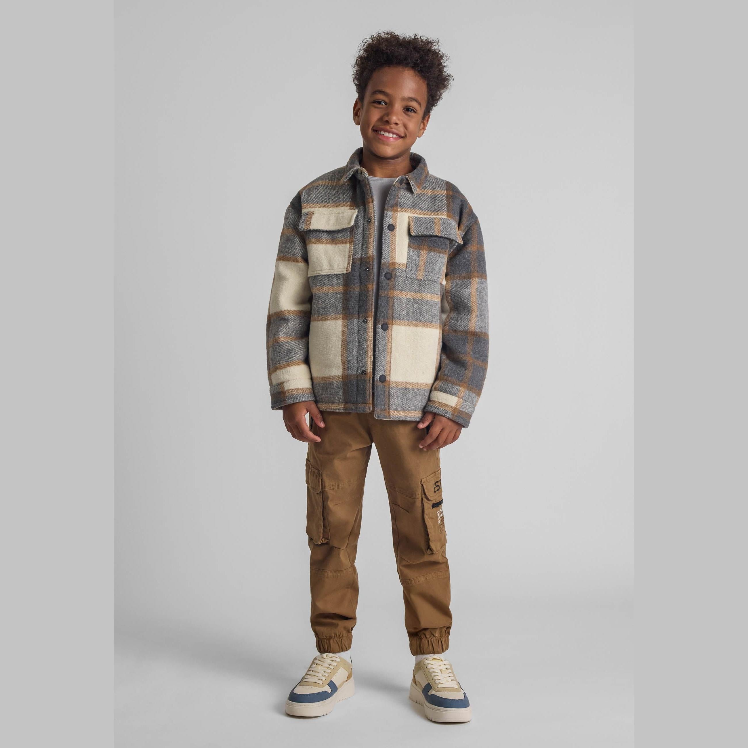 Young boy wearing beige cargo trousers with plaid shirt jacket, standing against white background, full body view showing relaxed casual styling
