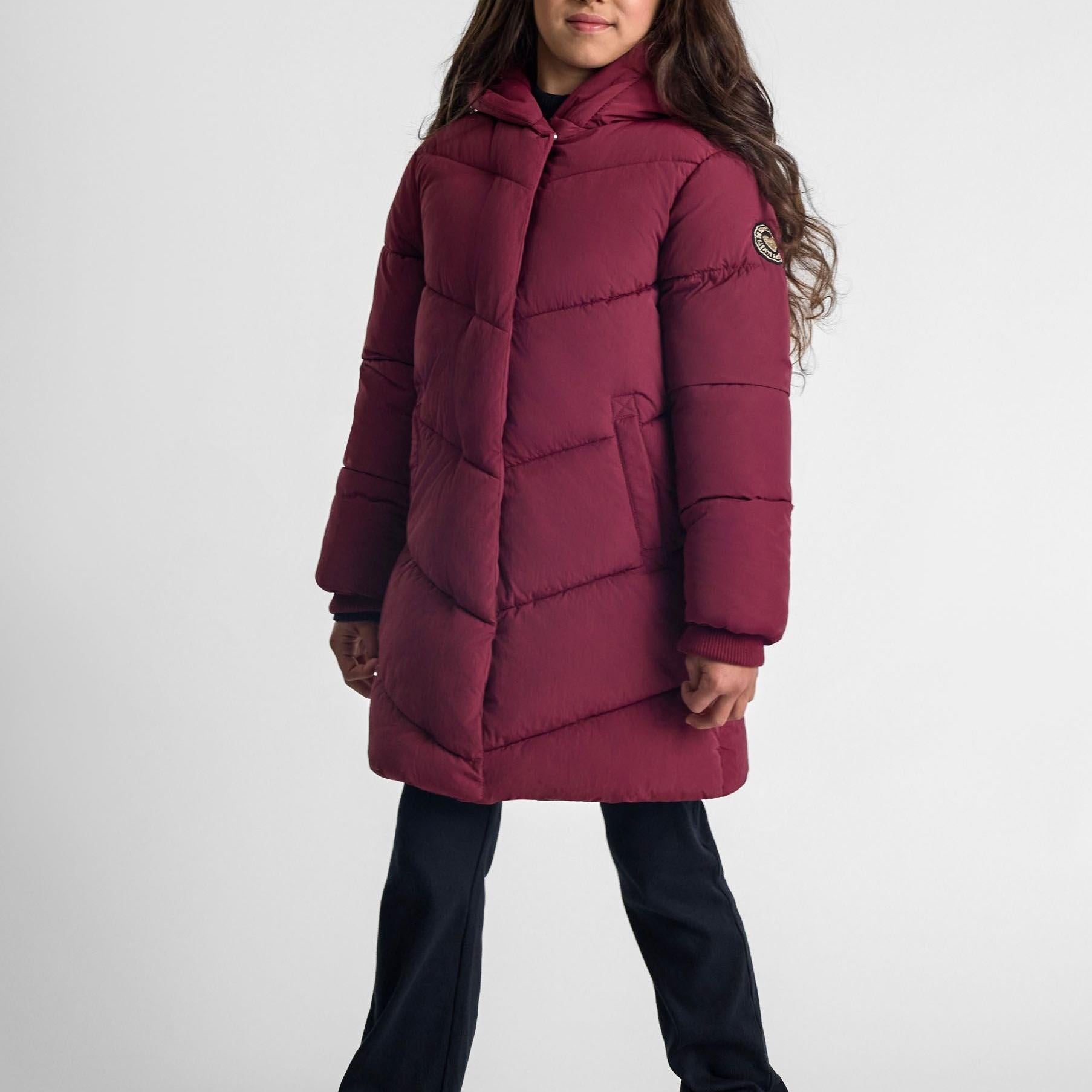 Girl wearing burgundy quilted winter jacket with hood and long sleeves, paired with dark jeans against grey background