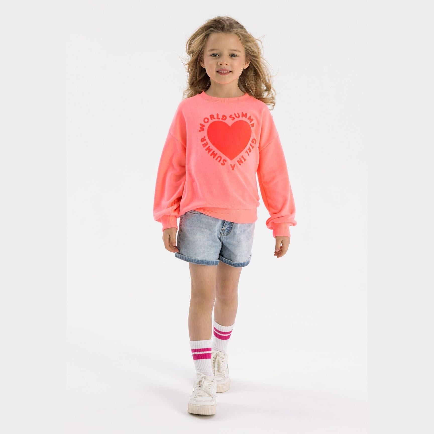 Young blonde girl wearing coral crew neck sweatshirt with heart print, denim shorts and white sneakers, standing against white background