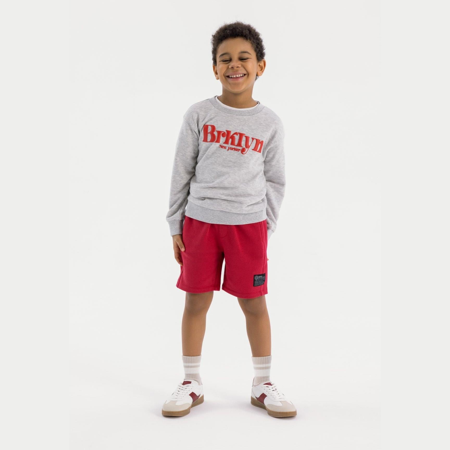 Young boy wearing grey sweatshirt with red text and red fleece shorts, smiling against white background, full body view showing complete casual outfit