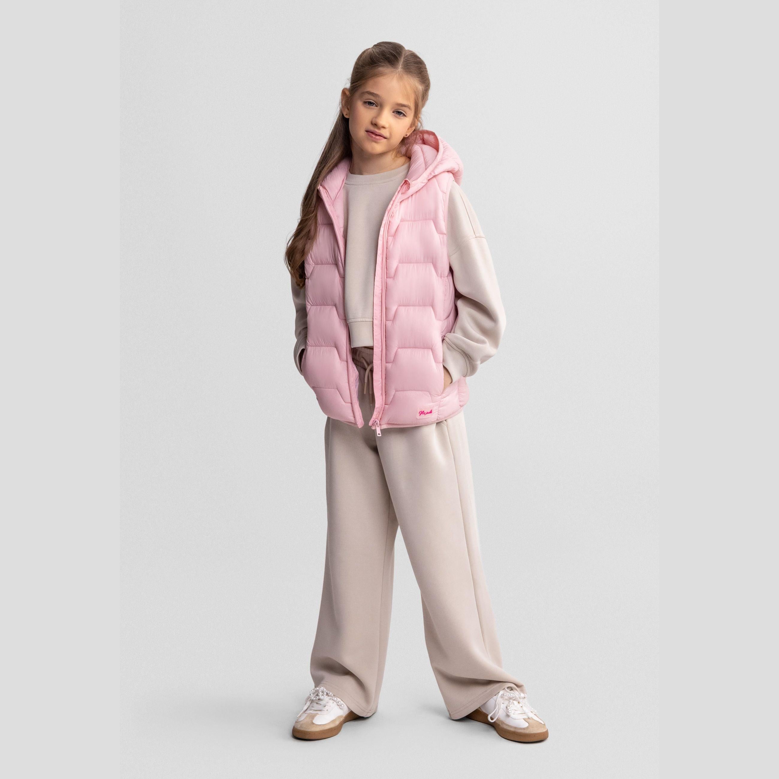 Young girl wearing pink quilted gilet over beige tracksuit standing against light grey background