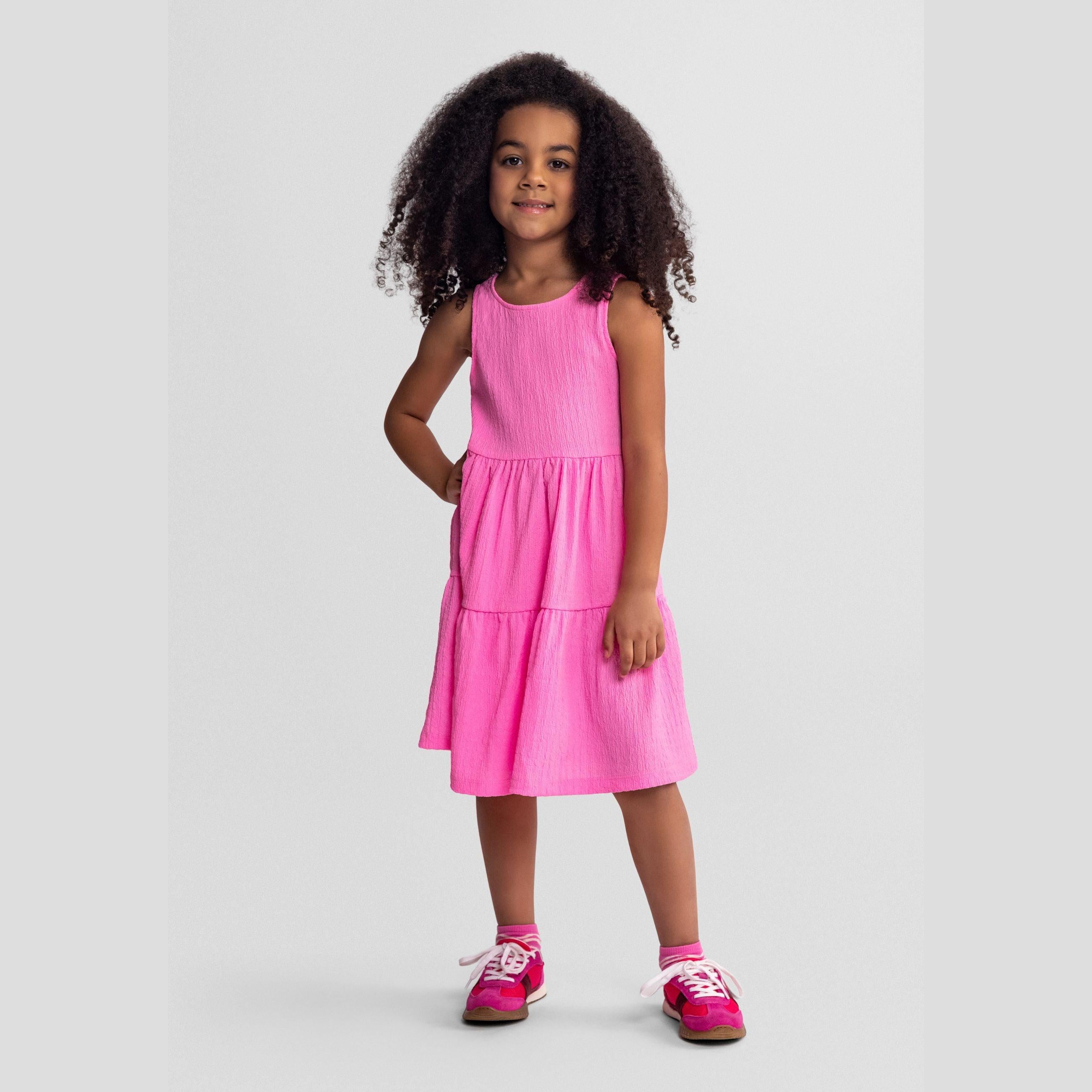 Young girl wearing bright pink tiered sleeveless dress with curly hair, standing against light grey background, full body front view
