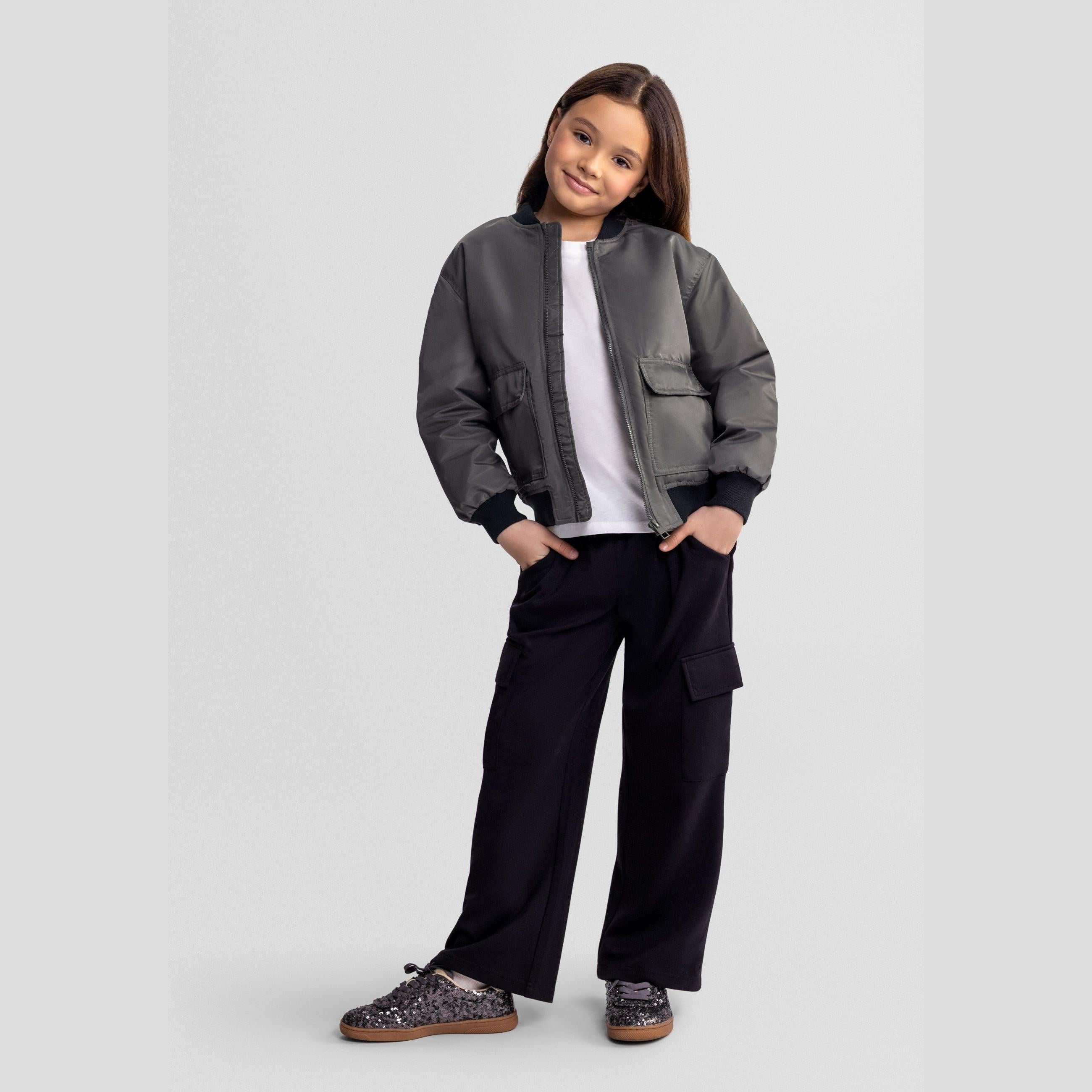 Young girl wearing black wide leg joggers with grey bomber jacket and white t-shirt, posing against light grey background with hands in pockets