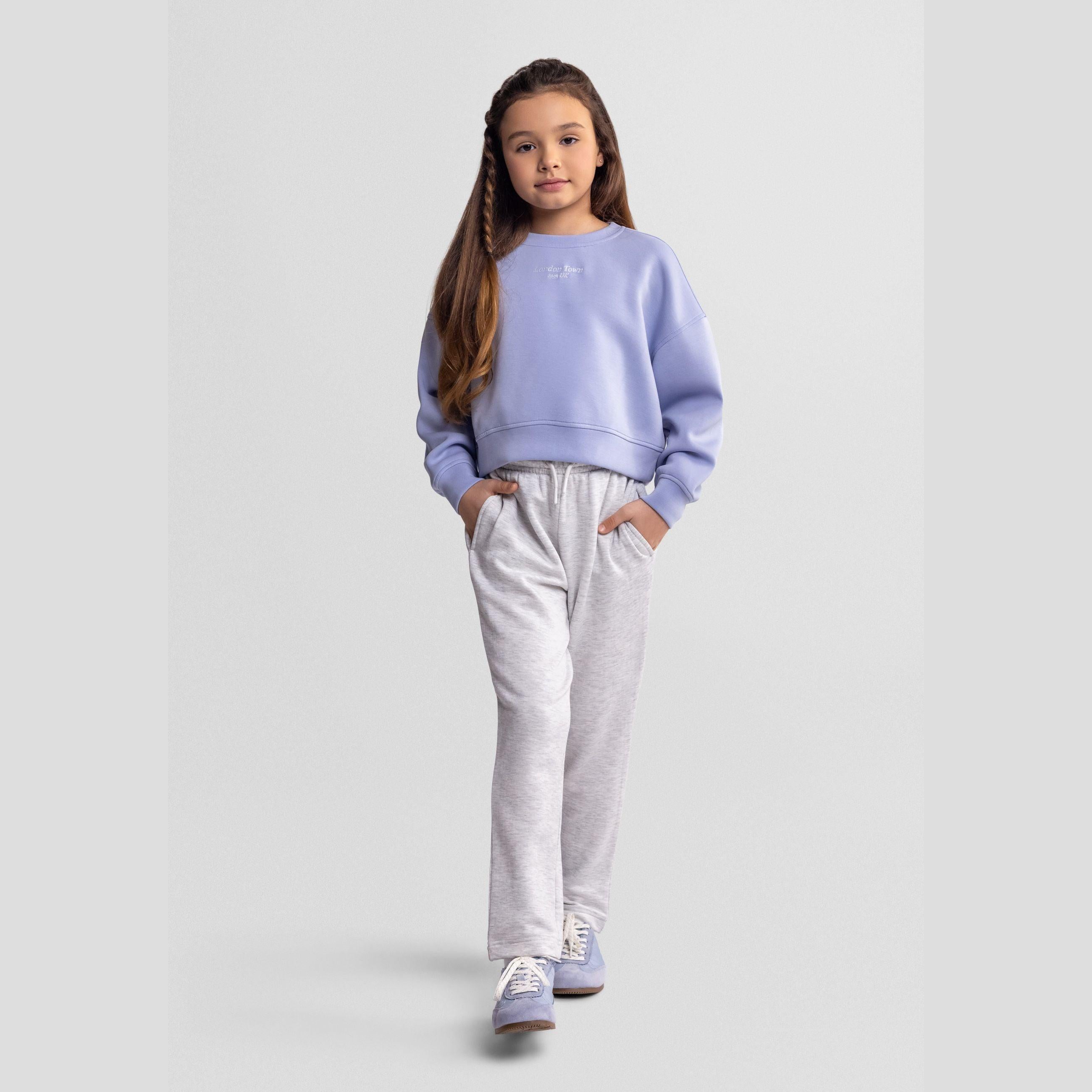 Young girl wearing light grey wide leg joggers with purple sweatshirt, standing against white background in casual pose