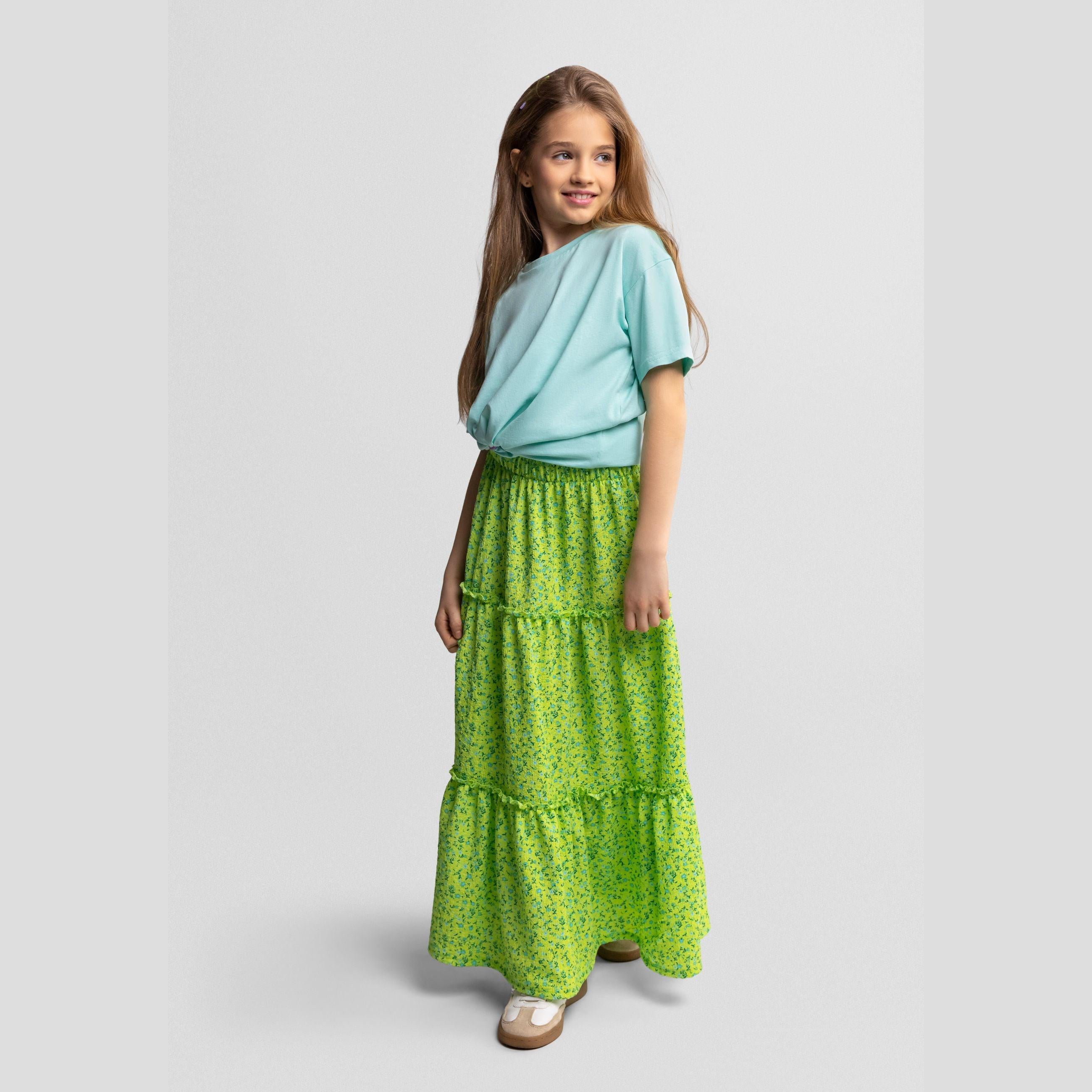 Young girl wearing mint green t-shirt and green floral maxi skirt with tiered ruffles, standing against light grey background