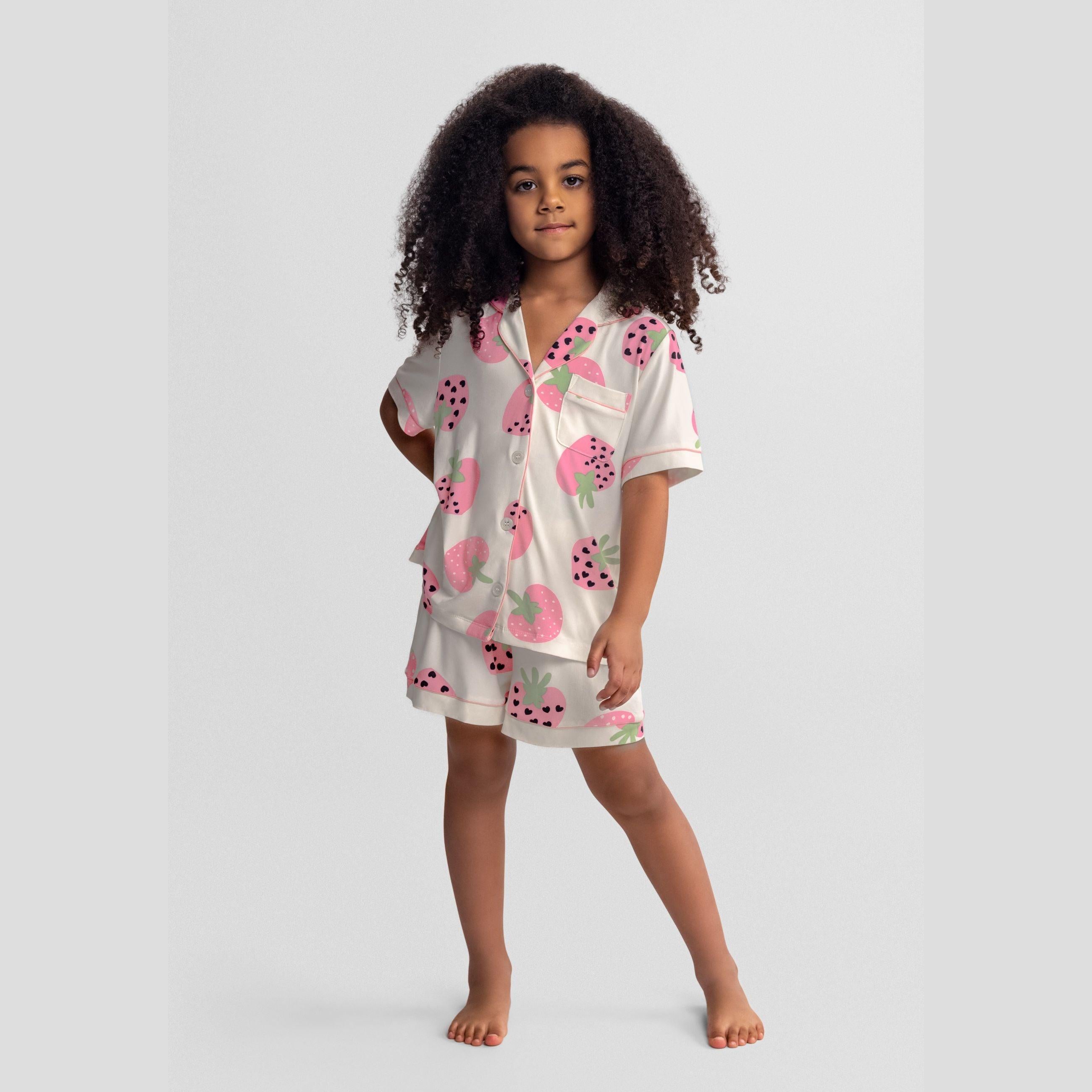 Girl wearing cream pyjama set with pink strawberry print, short sleeve top and matching shorts, standing against grey background