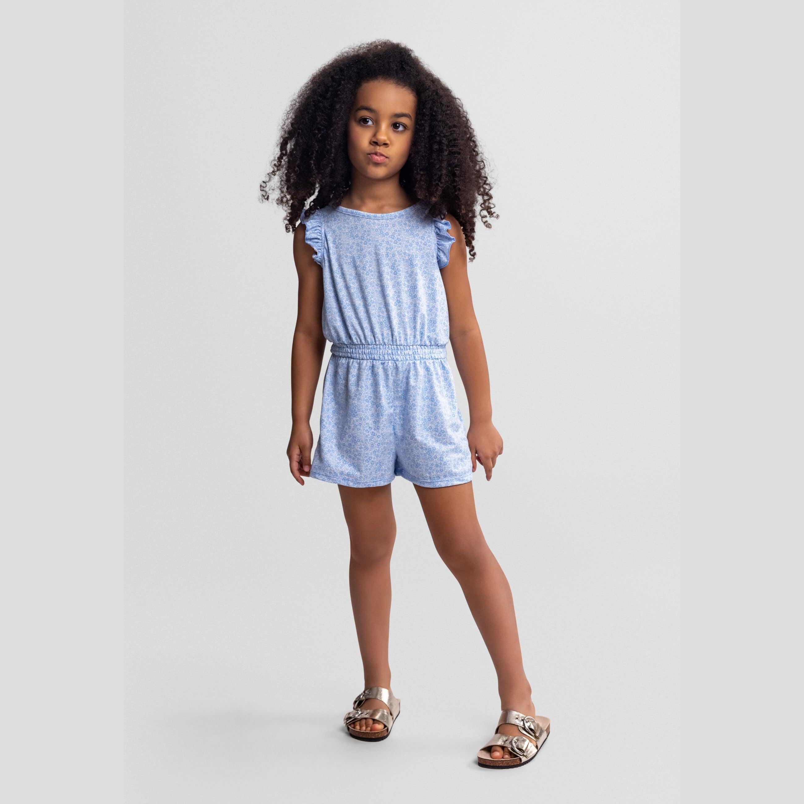 Young girl wearing blue floral jumpsuit with ruffle sleeves against light grey background, full body front view