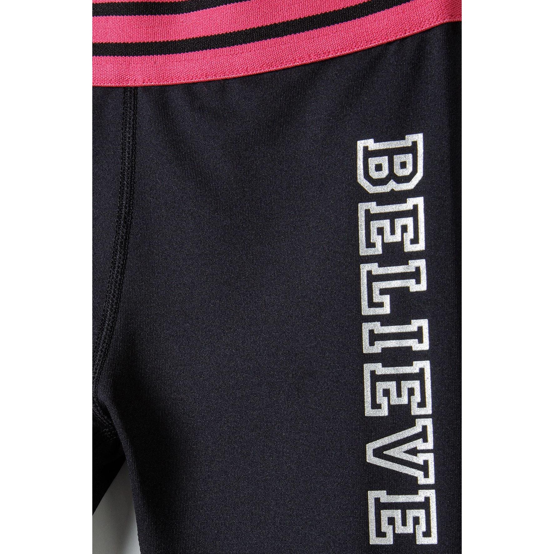 Close-up detail of black leggings showing silver foil BELIEVE text printed vertically down the leg with pink striped waistband visible at top