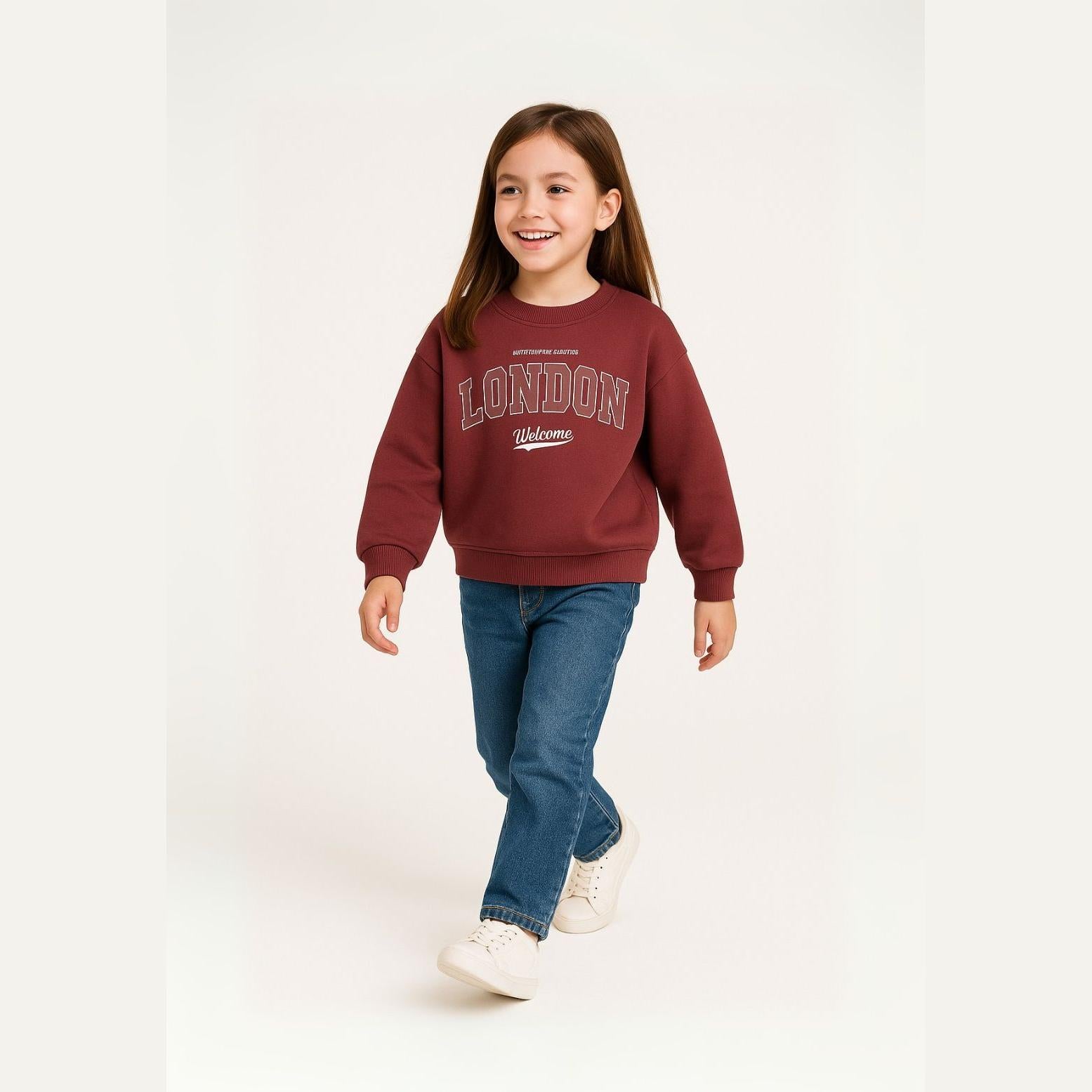 Young girl wearing burgundy crew sweatshirt with London print, paired with blue jeans and white sneakers, walking pose against light background
