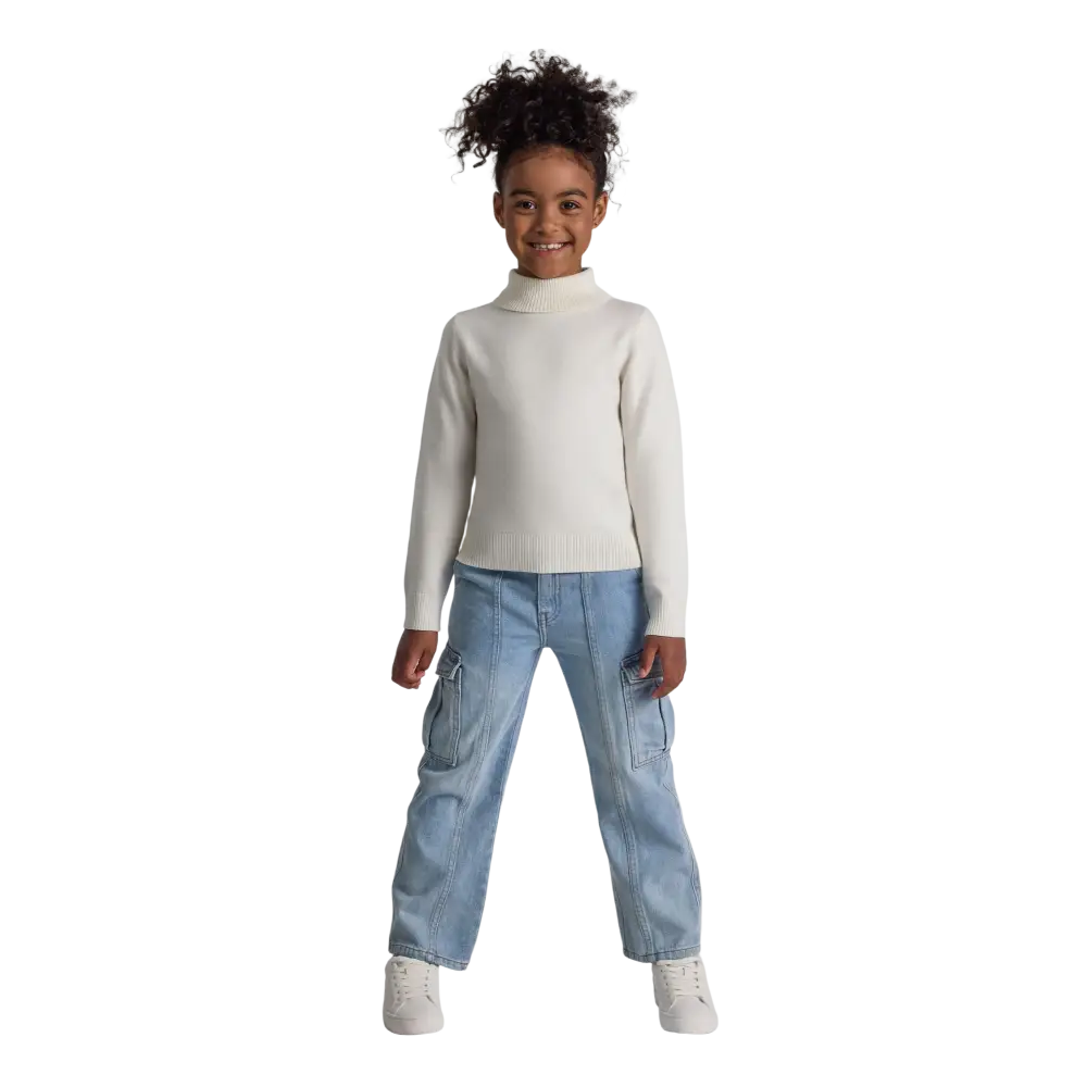 Young girl wearing cream fine knit turtleneck jumper with light blue cargo pants and white sneakers, standing against black background