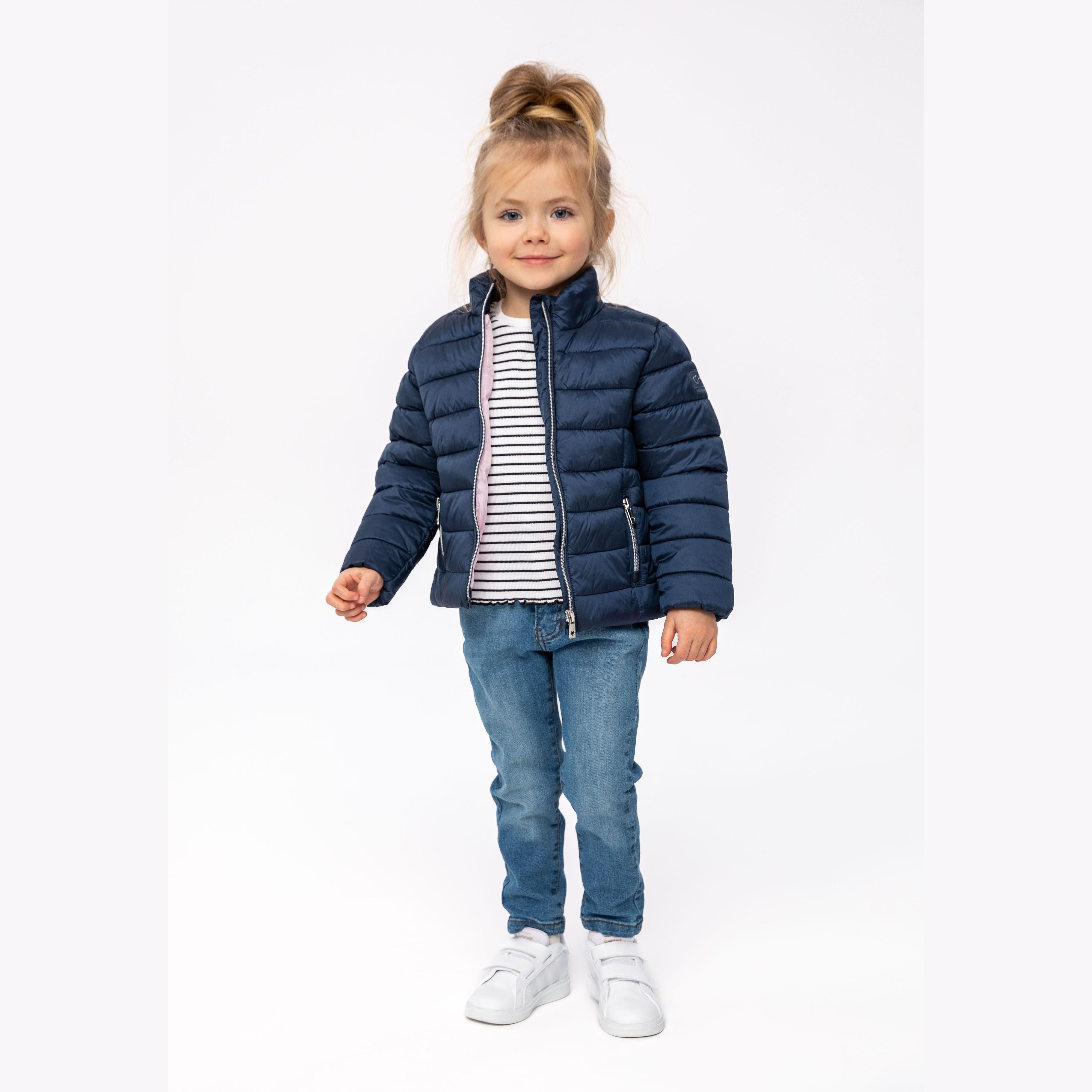 Young blonde girl wearing navy quilted padded jacket over striped top with jeans and white trainers, standing against white background