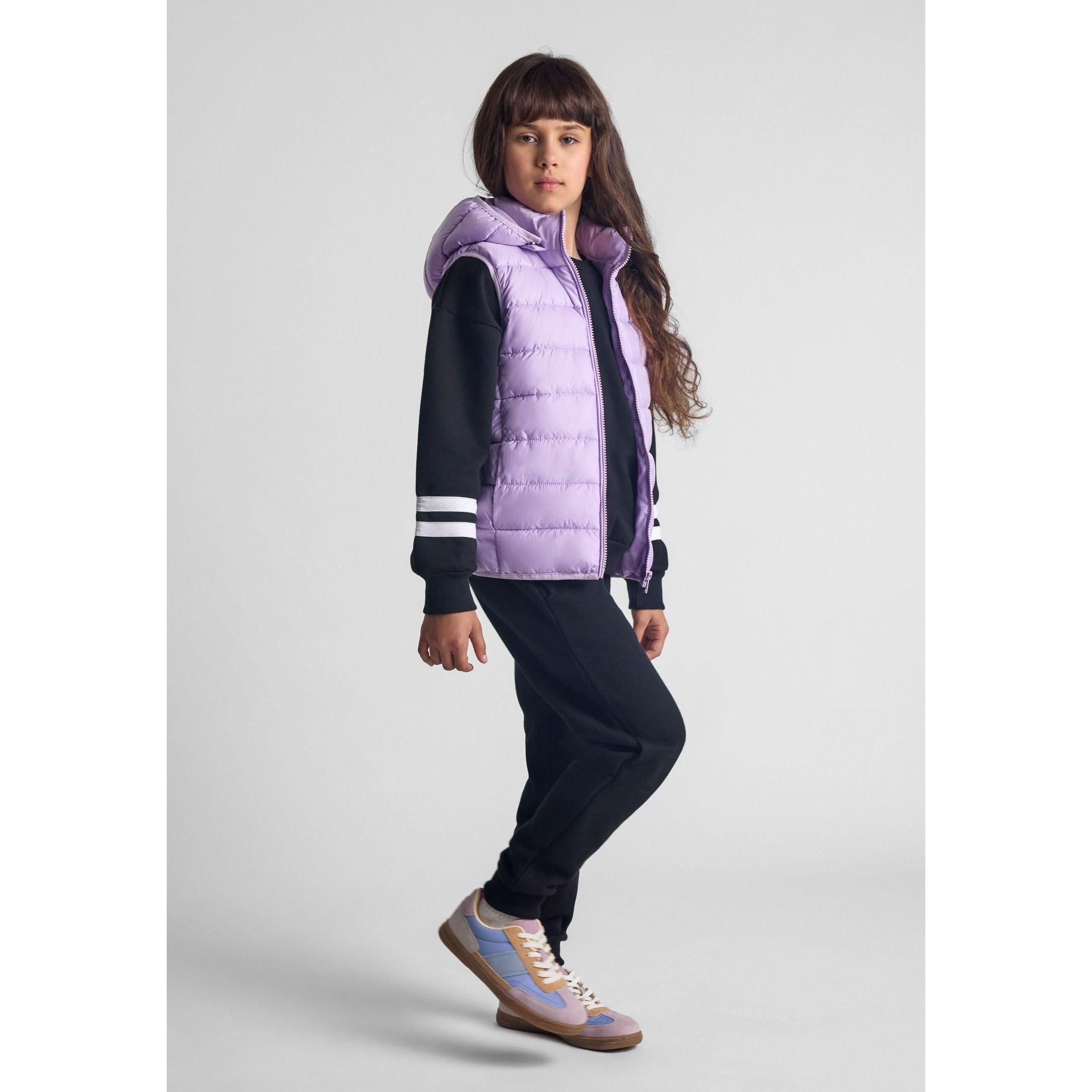 Young girl wearing lilac quilted gilet over navy tracksuit with white stripes, standing against grey background