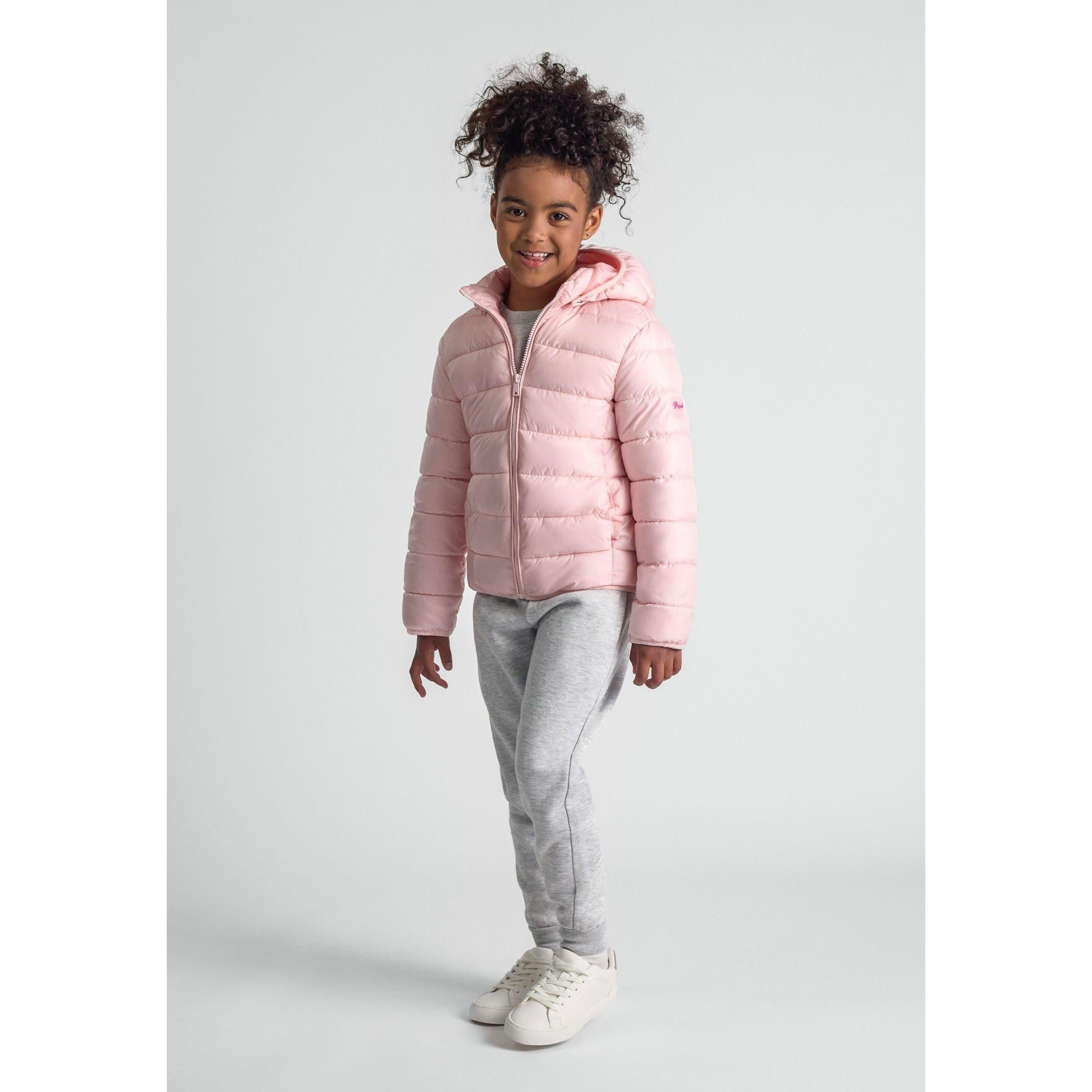 Young girl wearing pink quilted jacket with detachable hood and grey joggers, standing against light grey background