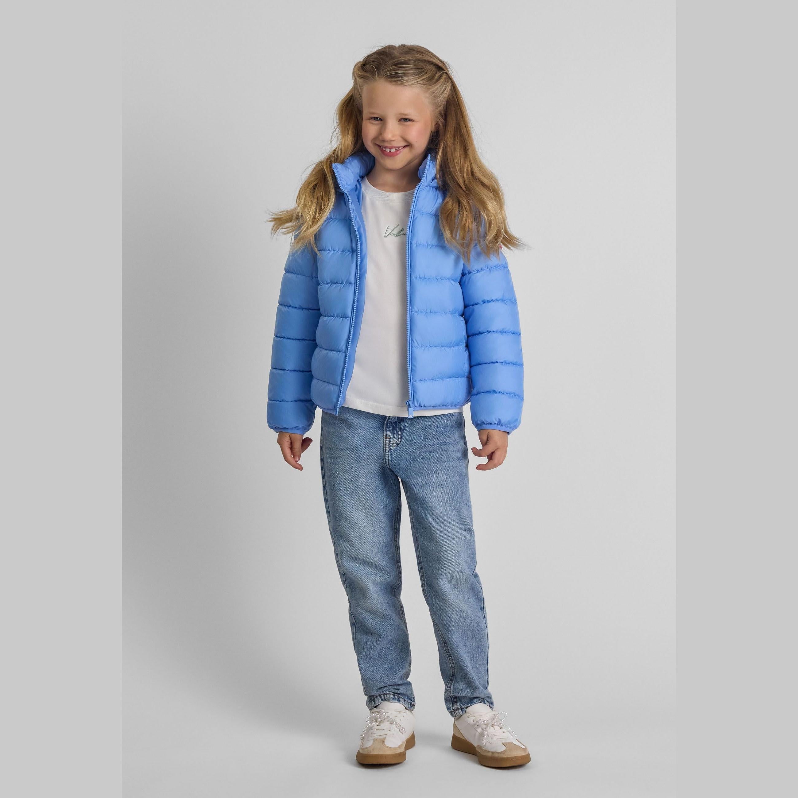 Young girl wearing blue quilted jacket with detachable hood, white top and jeans, smiling against light grey background