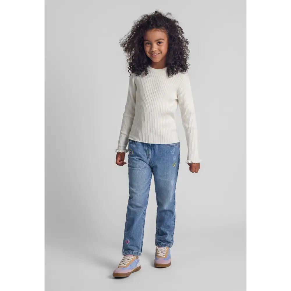 Young model wearing white ribbed jumper with frill cuffs paired with blue jeans, full body shot on light grey background